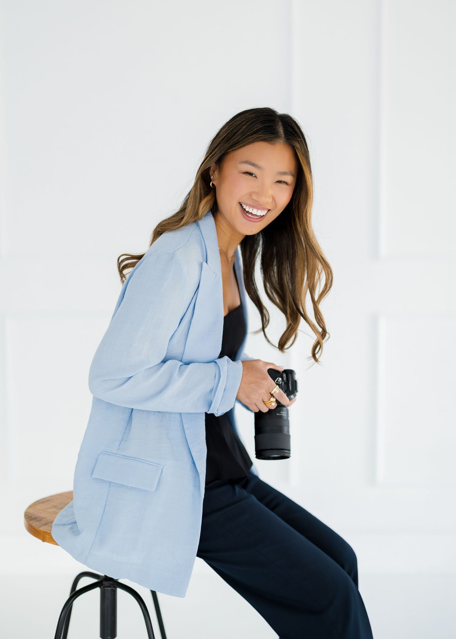 Woman in light blue blazer and dark pants sitting casually in white studio setting.