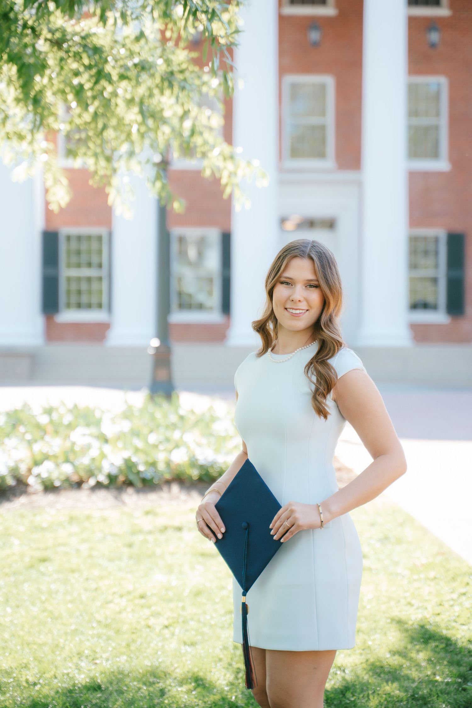 Person in white dress holding graduation materials near classical building columns.