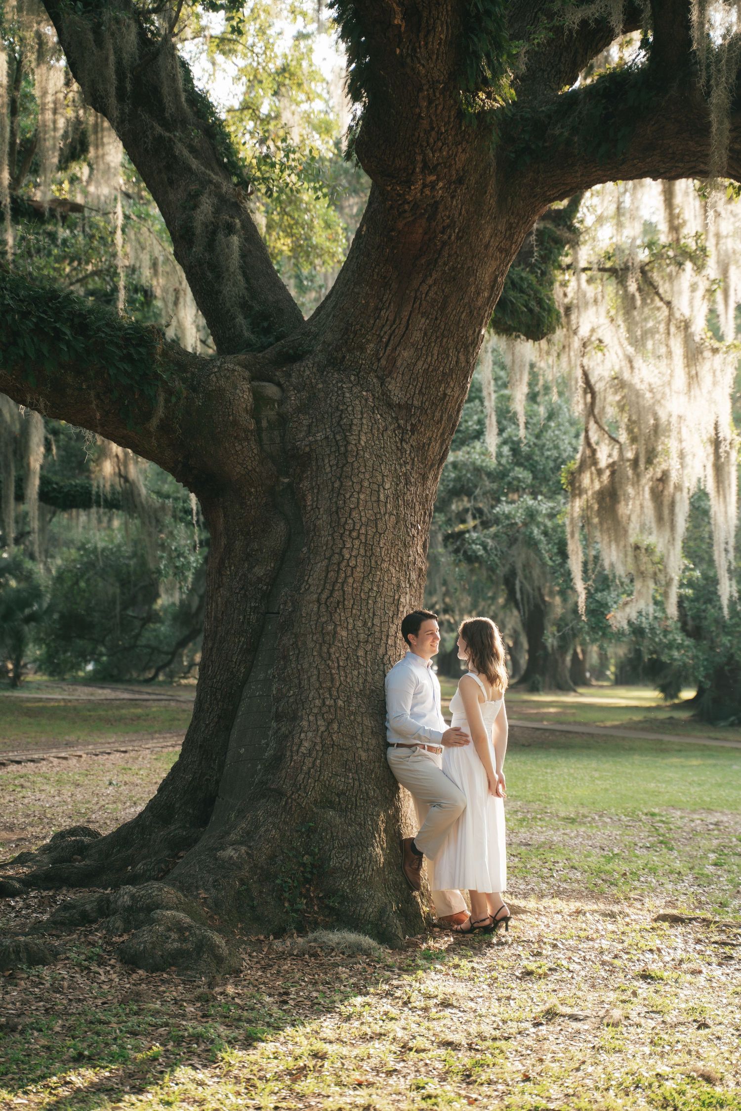 Couple sharing a romantic moment leaning against a massive oak tree draped in Spanish moss in a lush park.