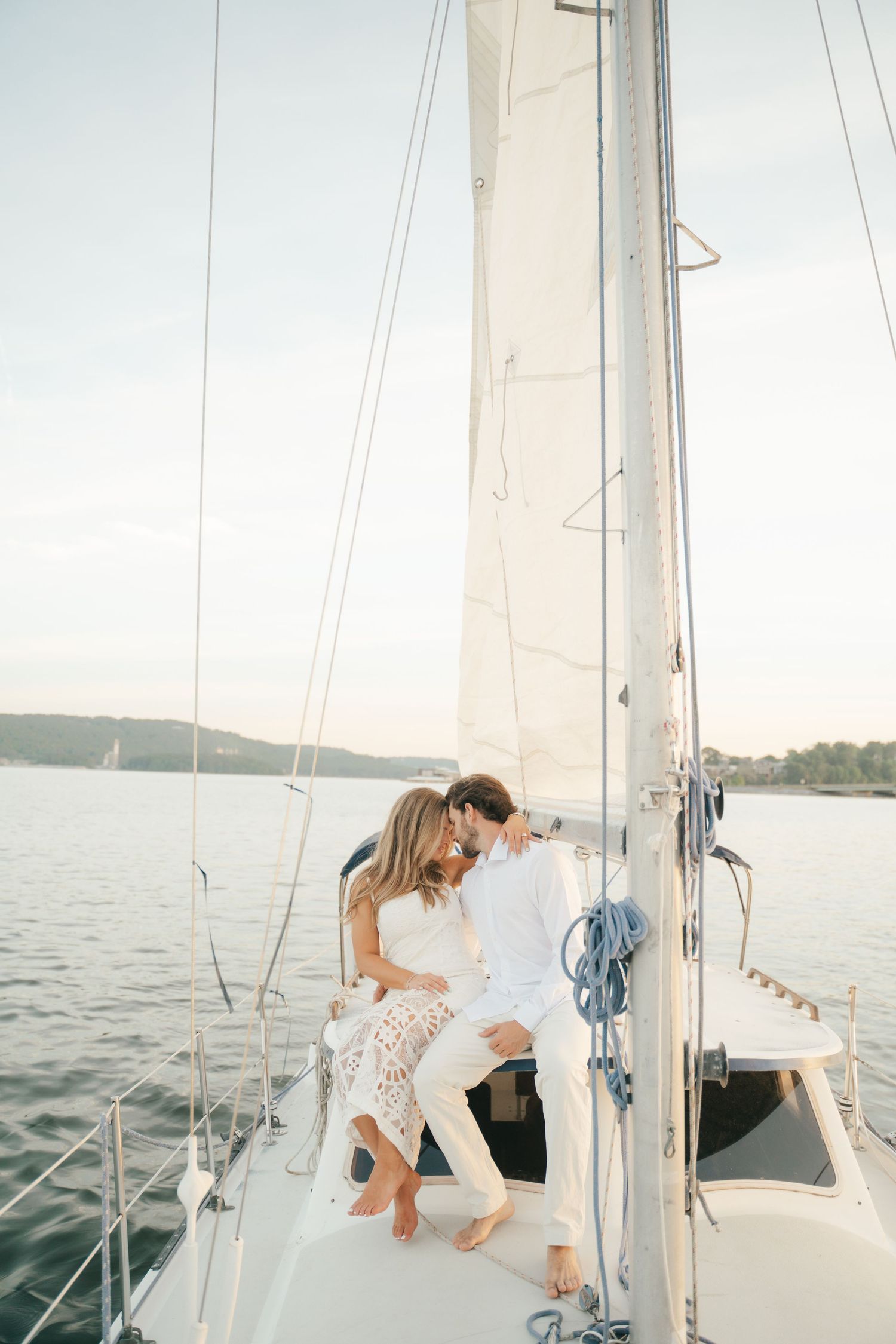 Romantic couple in white outfits sitting close together on a sailboat deck, with calm blue water and sky in the background.