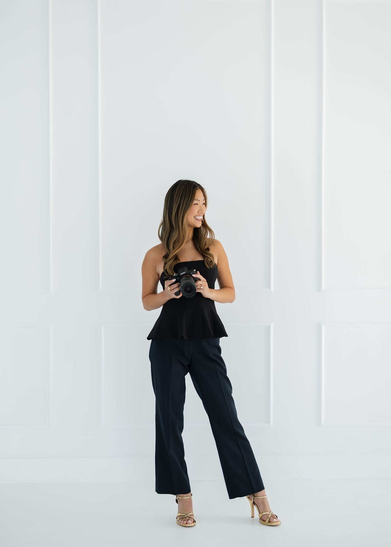 Woman in black strapless peplum top and trousers holding a camera, posing against a white paneled wall.