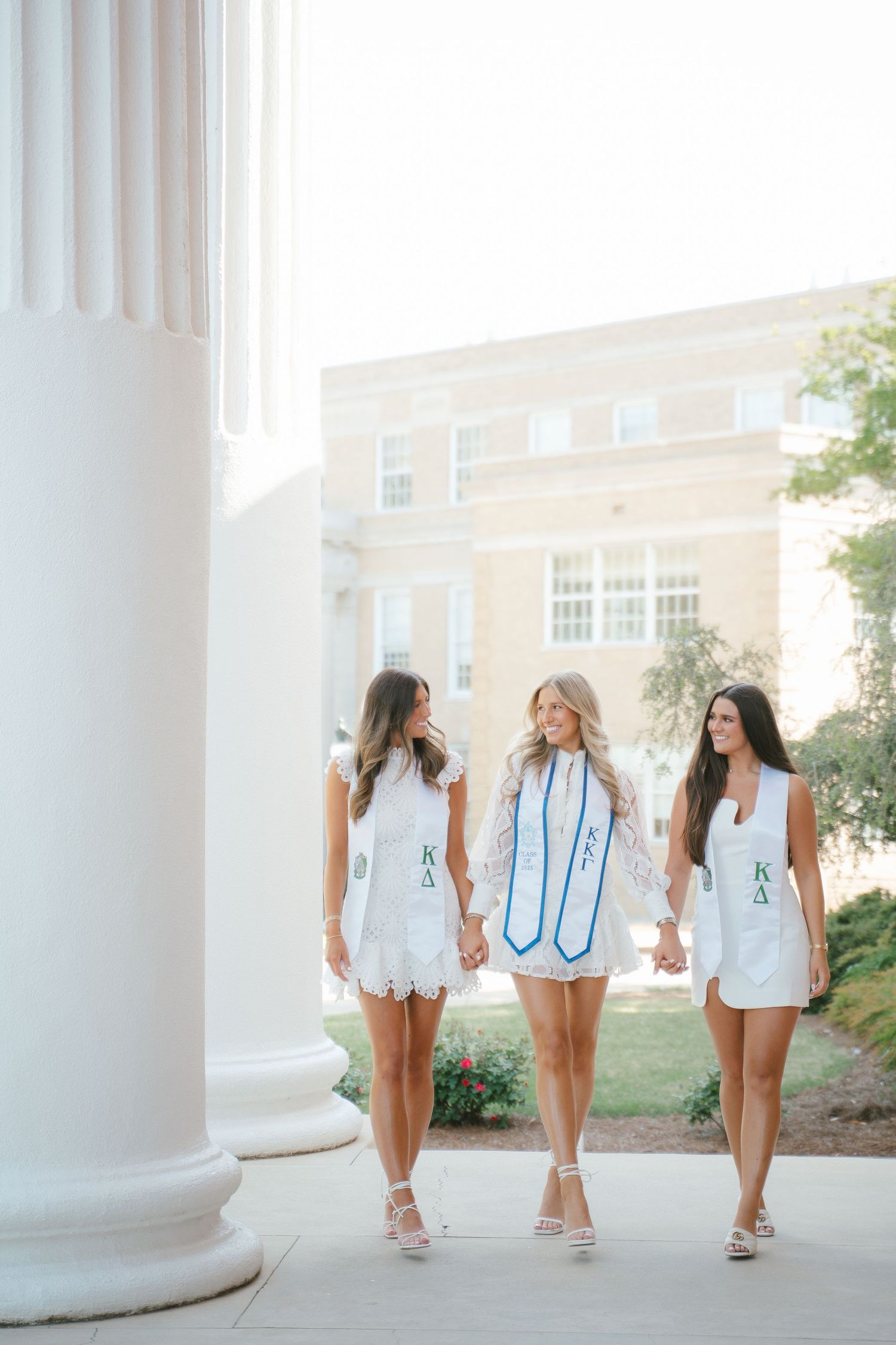 Three people in white outfits walk together between classical white columns on a sunny day.