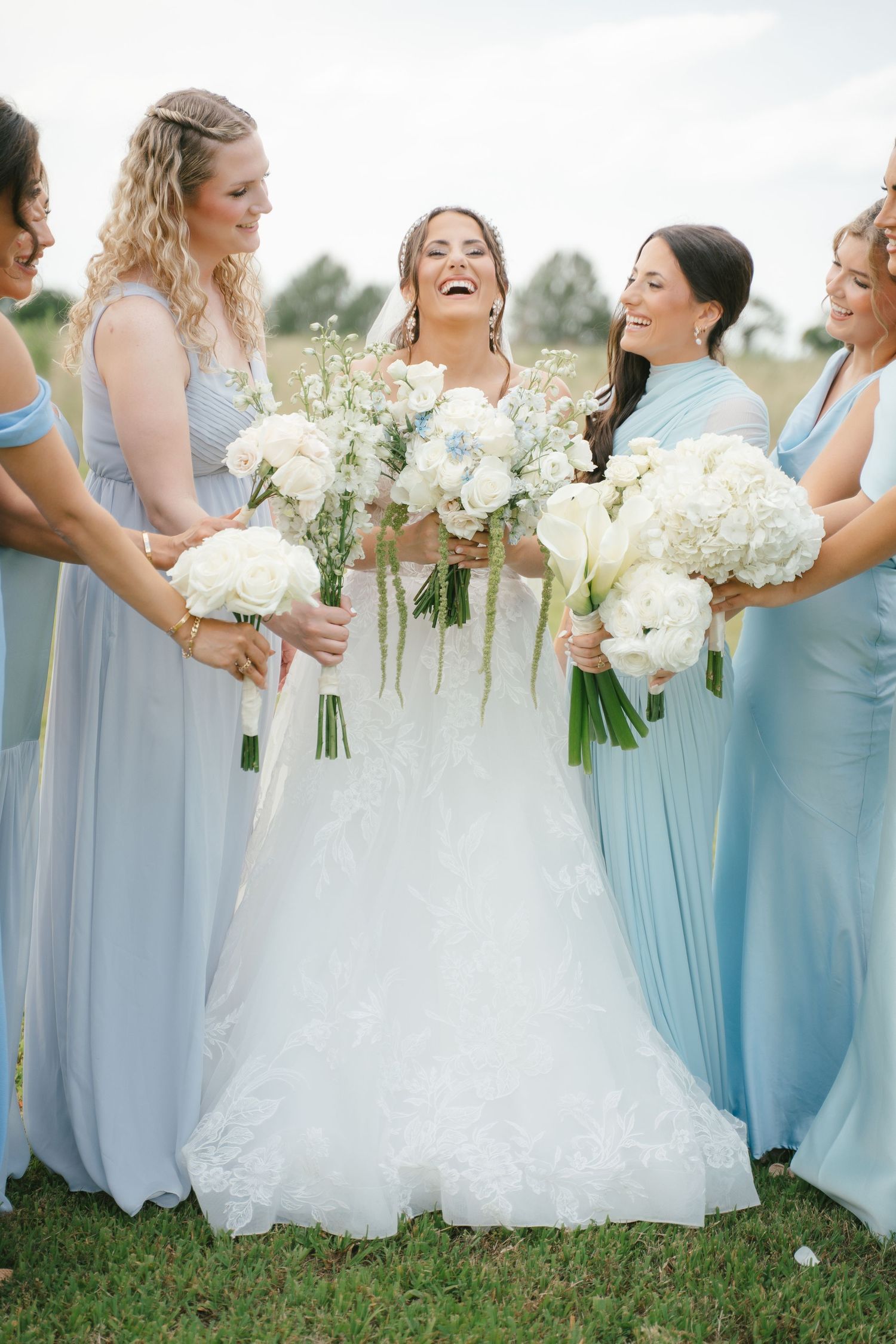 Bridesmaids in light blue dresses holding white bouquets pose together on grass.