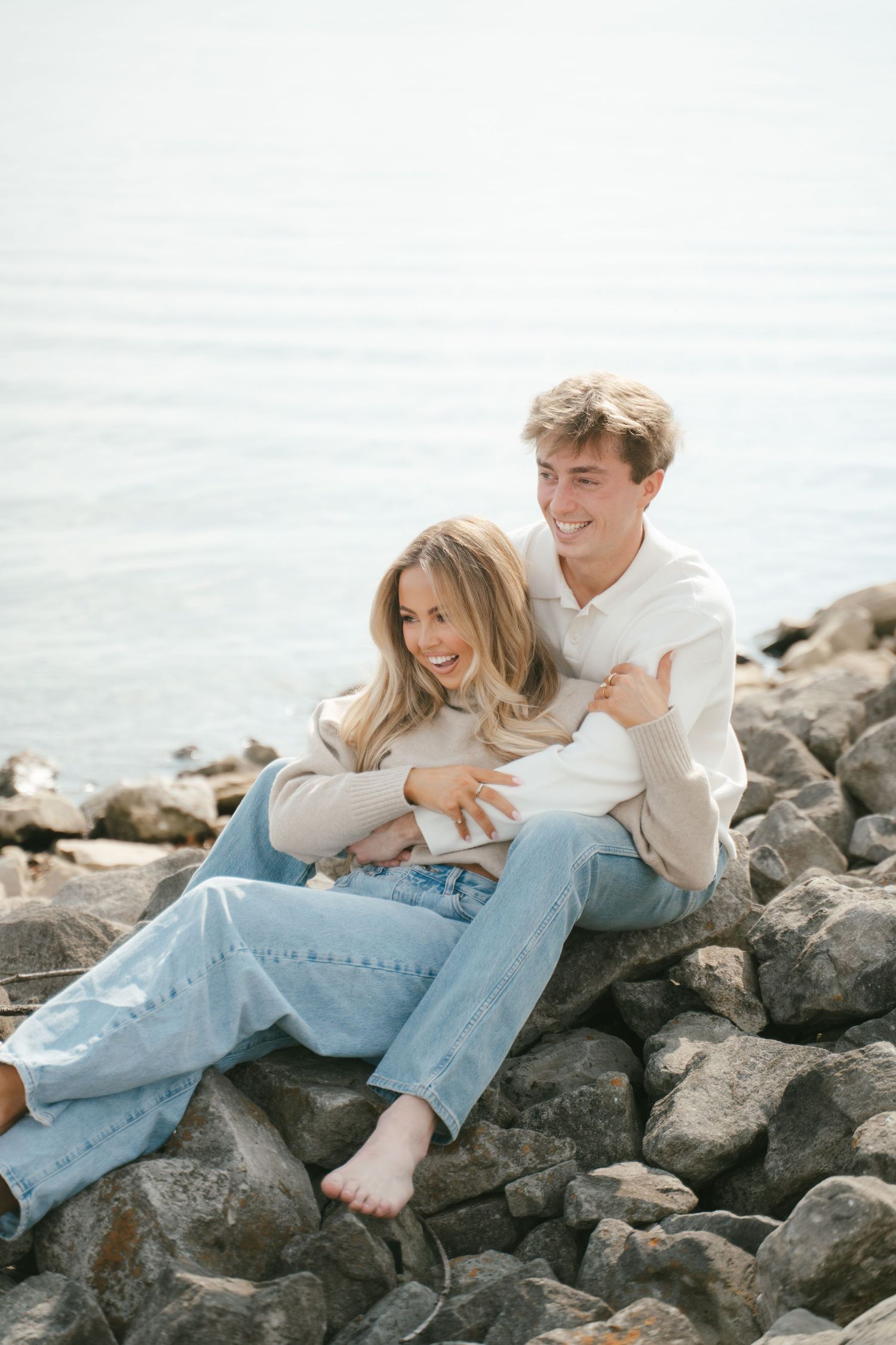 Couple embracing on rocky shoreline overlooking water.