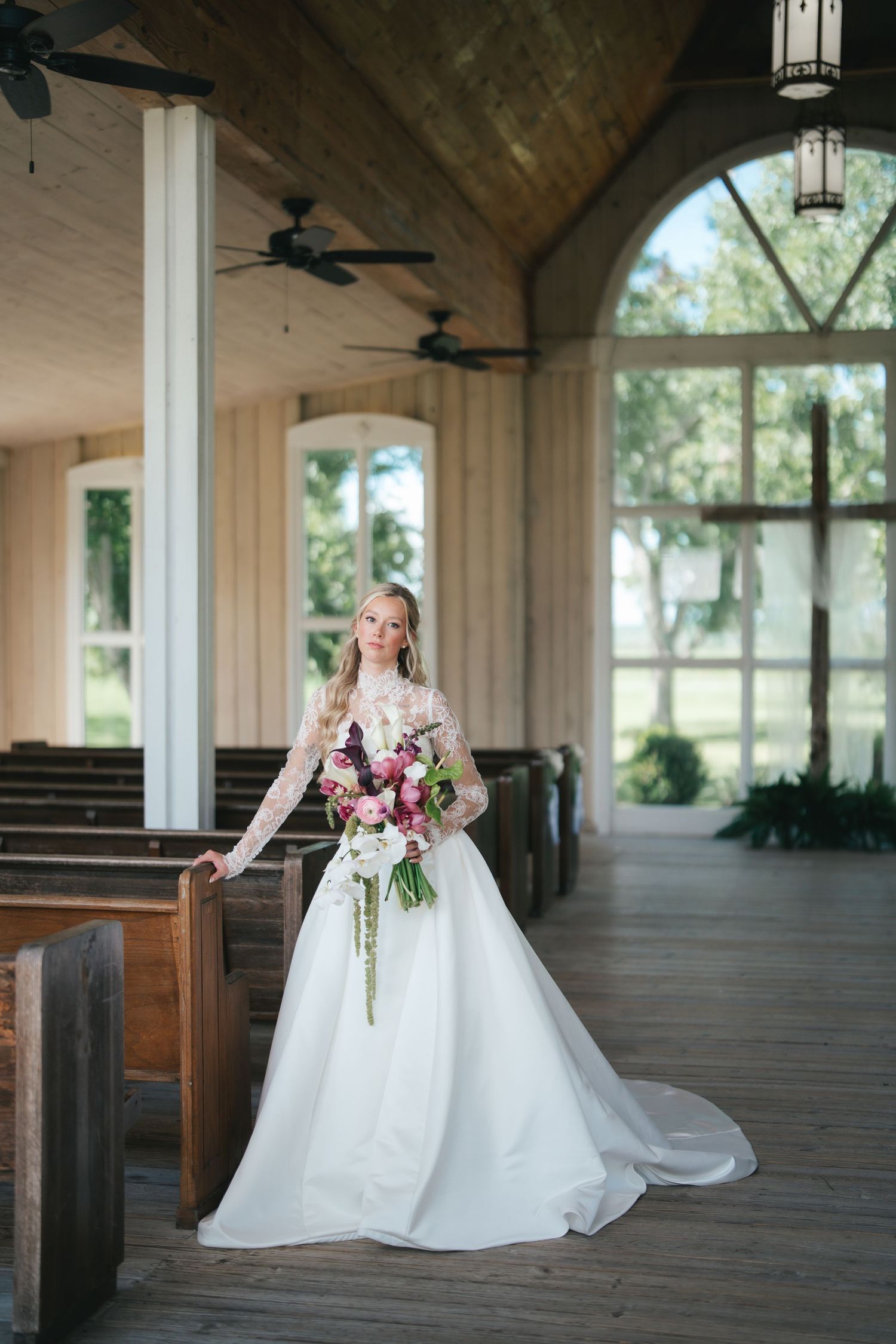 Bride in lace long-sleeve wedding gown holding floral bouquet standing in elegant wooden chapel with arched windows.
