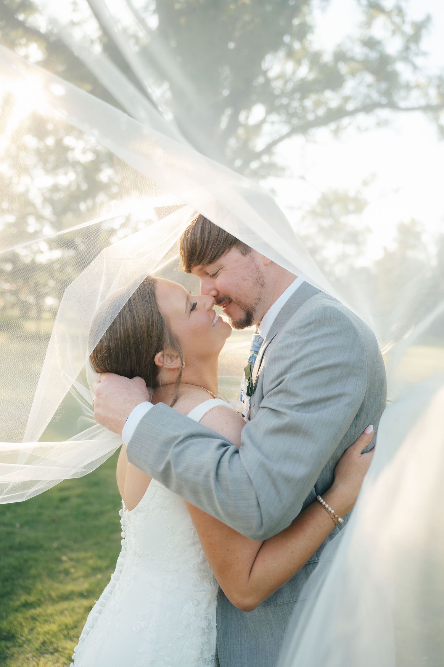 Bride and groom share a romantic moment under a flowing veil outdoors, with soft sunlight filtering through the trees.