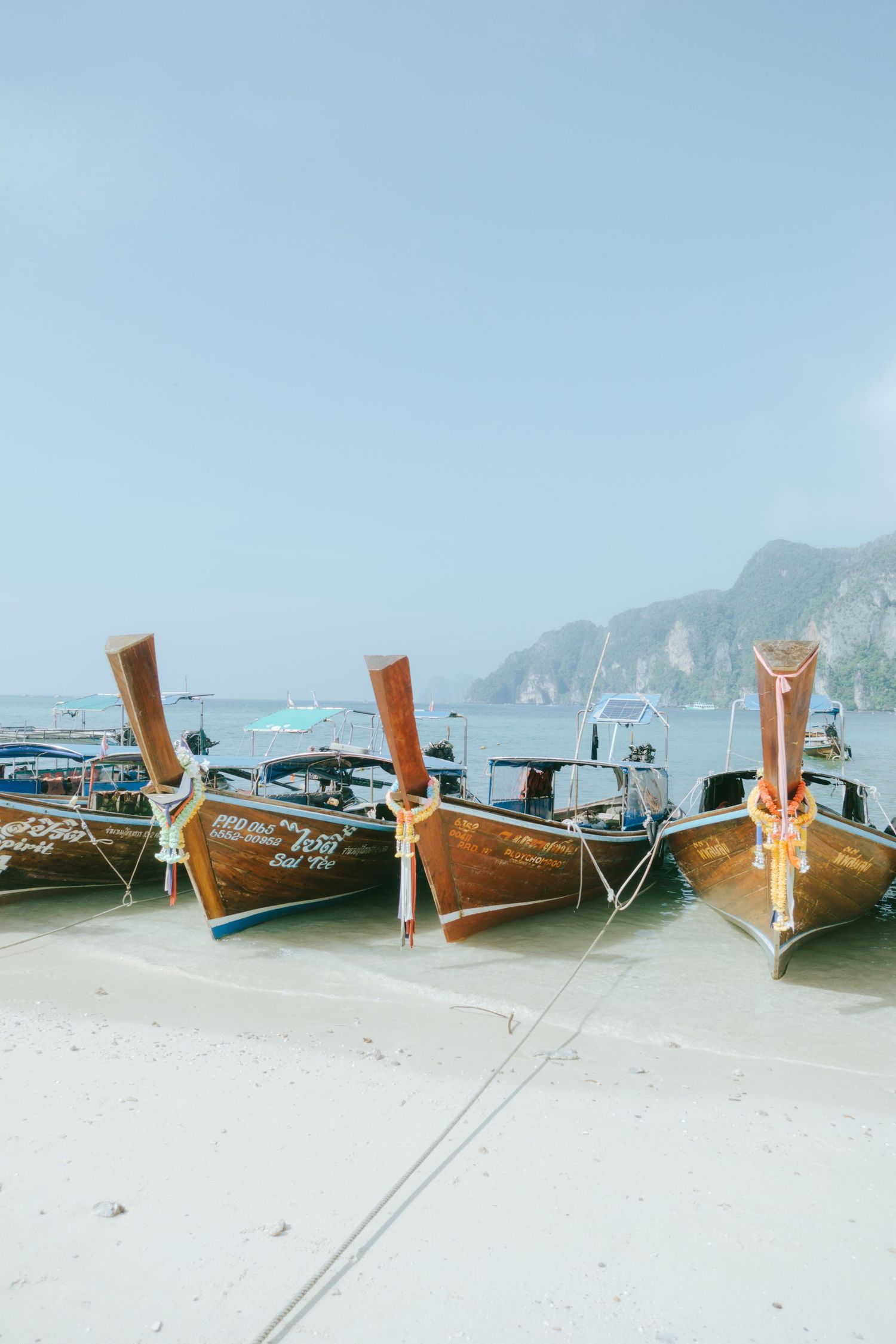 Traditional wooden longtail boats anchored on a white sandy beach with limestone cliffs in the background.