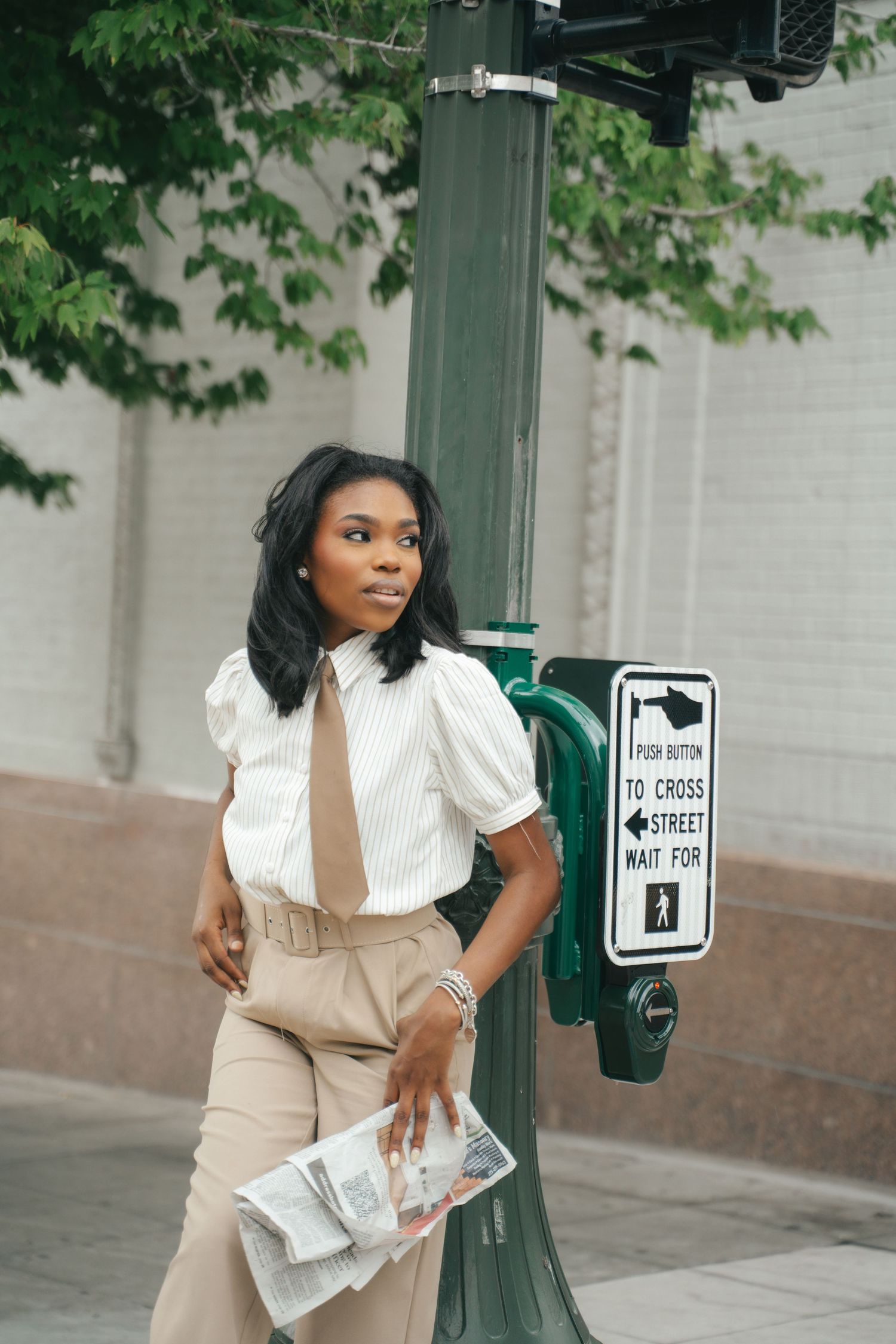 Stylish Black woman in white striped blouse and tan trousers poses by a street sign pole in an urban setting.