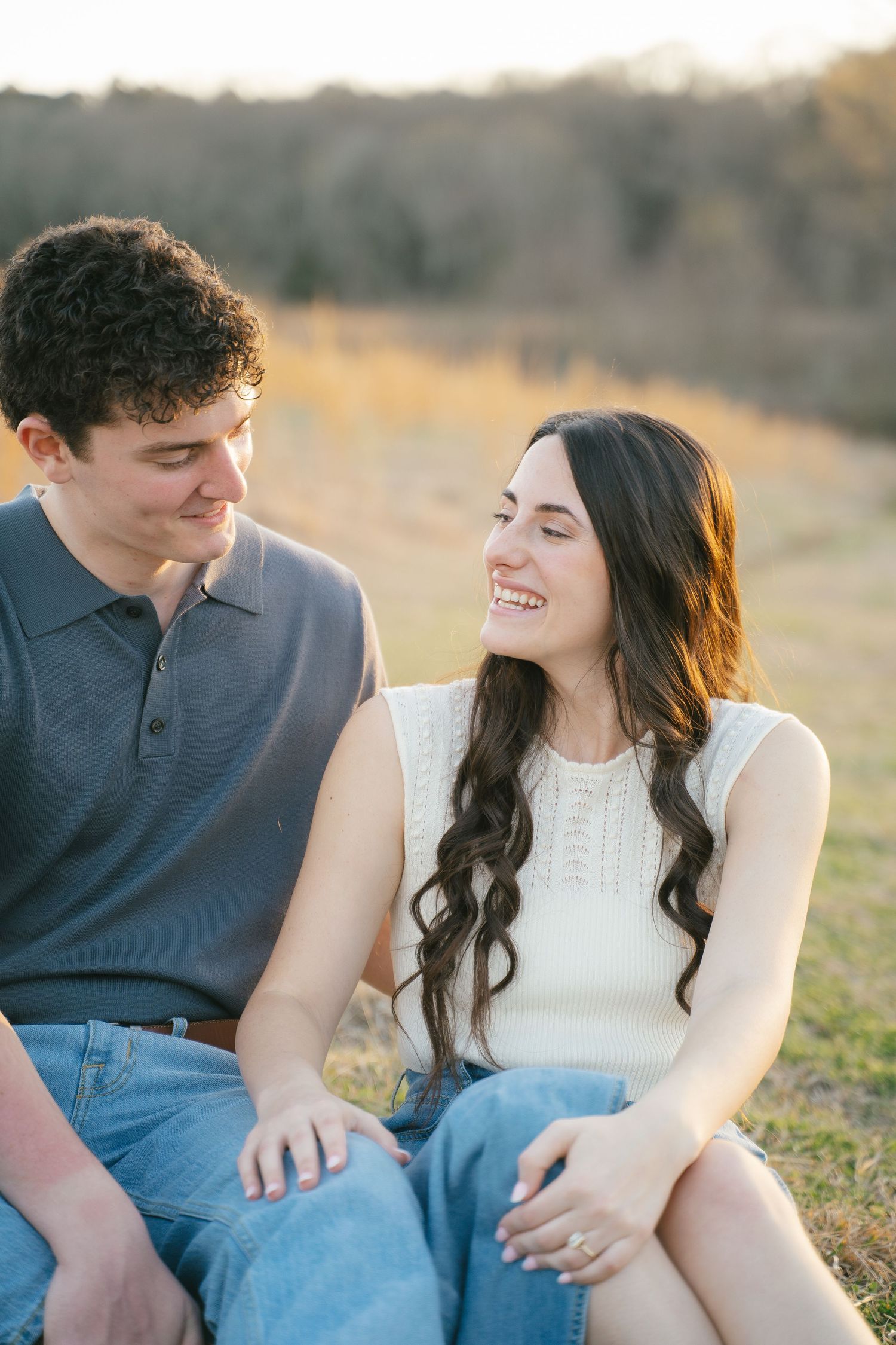 A couple laughs together while sitting in a grassy outdoor setting.