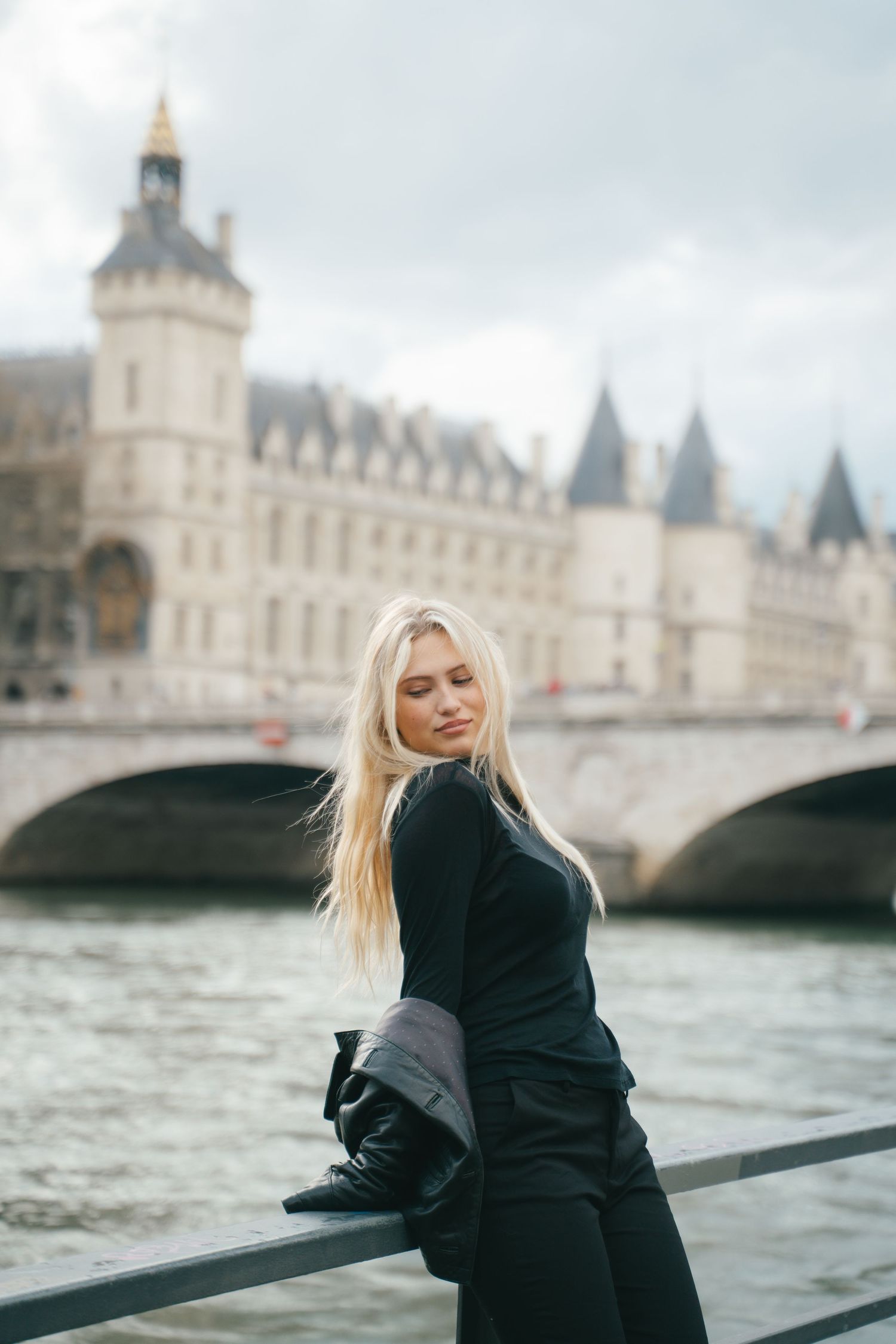 Blonde woman in black outfit posing by Seine River with Conciergerie castle and bridge in Paris background.