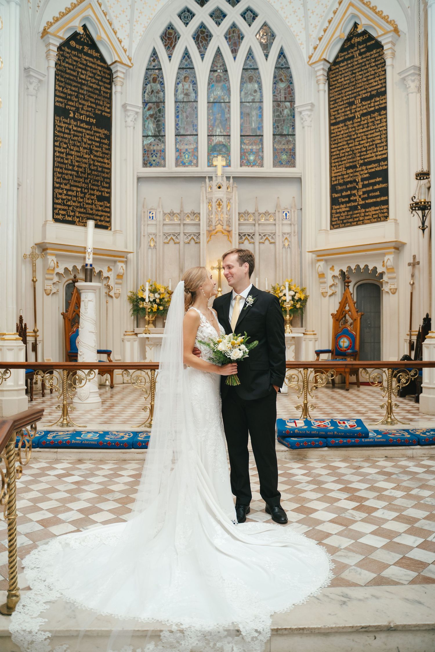 Bride and groom share a romantic moment at church altar, bride in elegant white gown with long train holding floral bouquet.