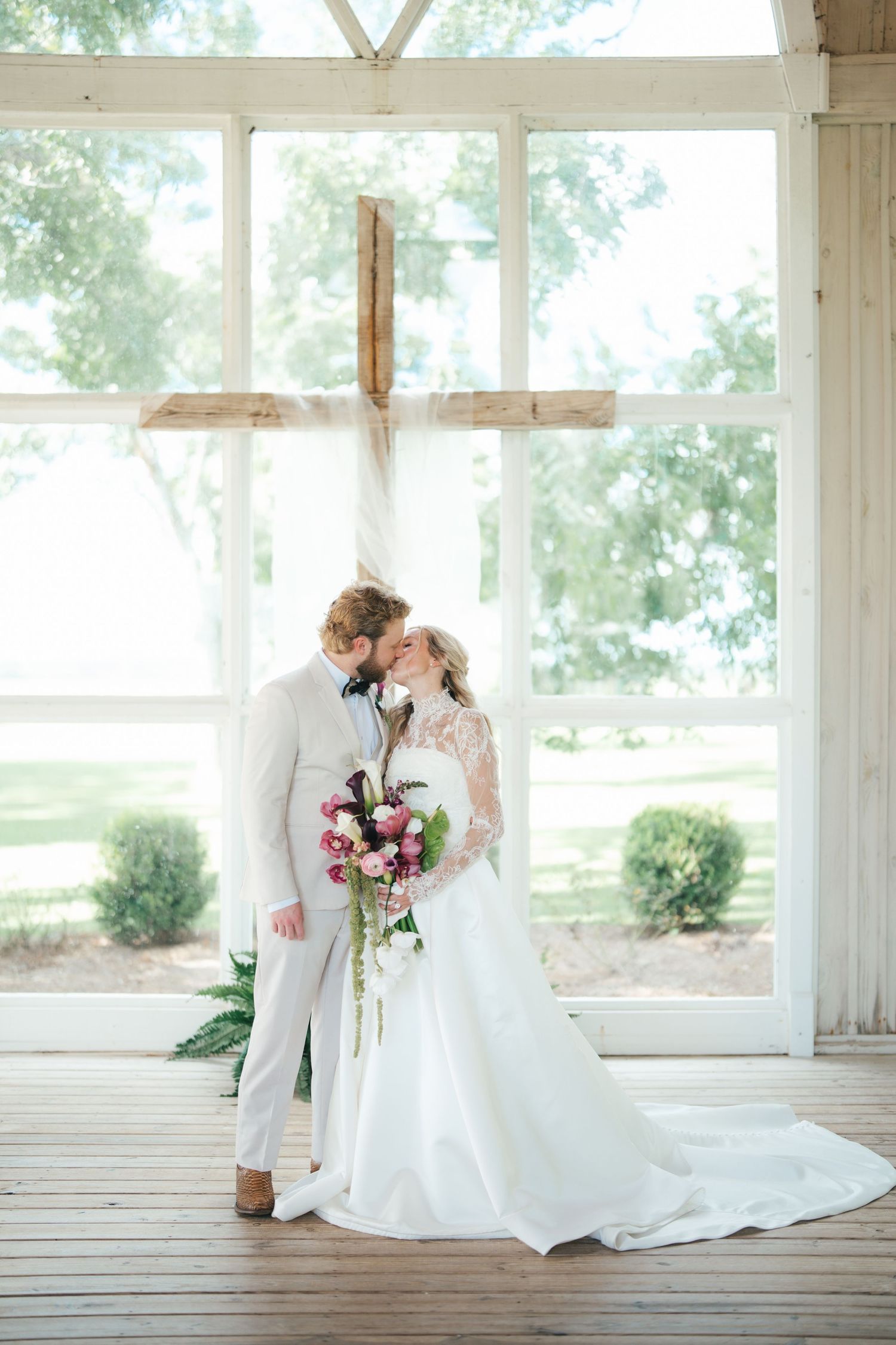Bride and groom share a kiss in a rustic chapel with a wooden cross and large windows overlooking lush greenery.