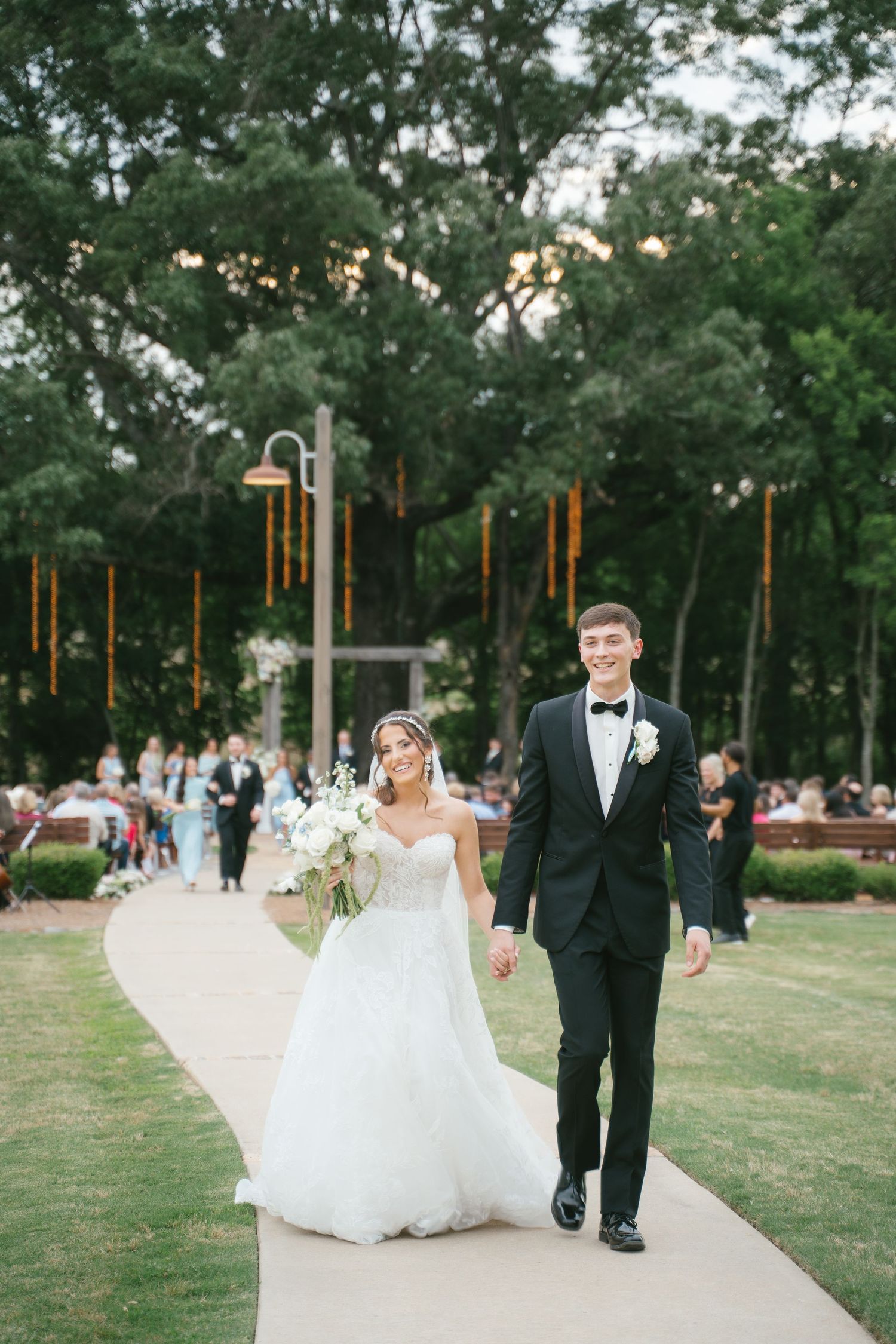 A bride and groom walk hand in hand down a garden path at dusk with string lights and trees in the background.