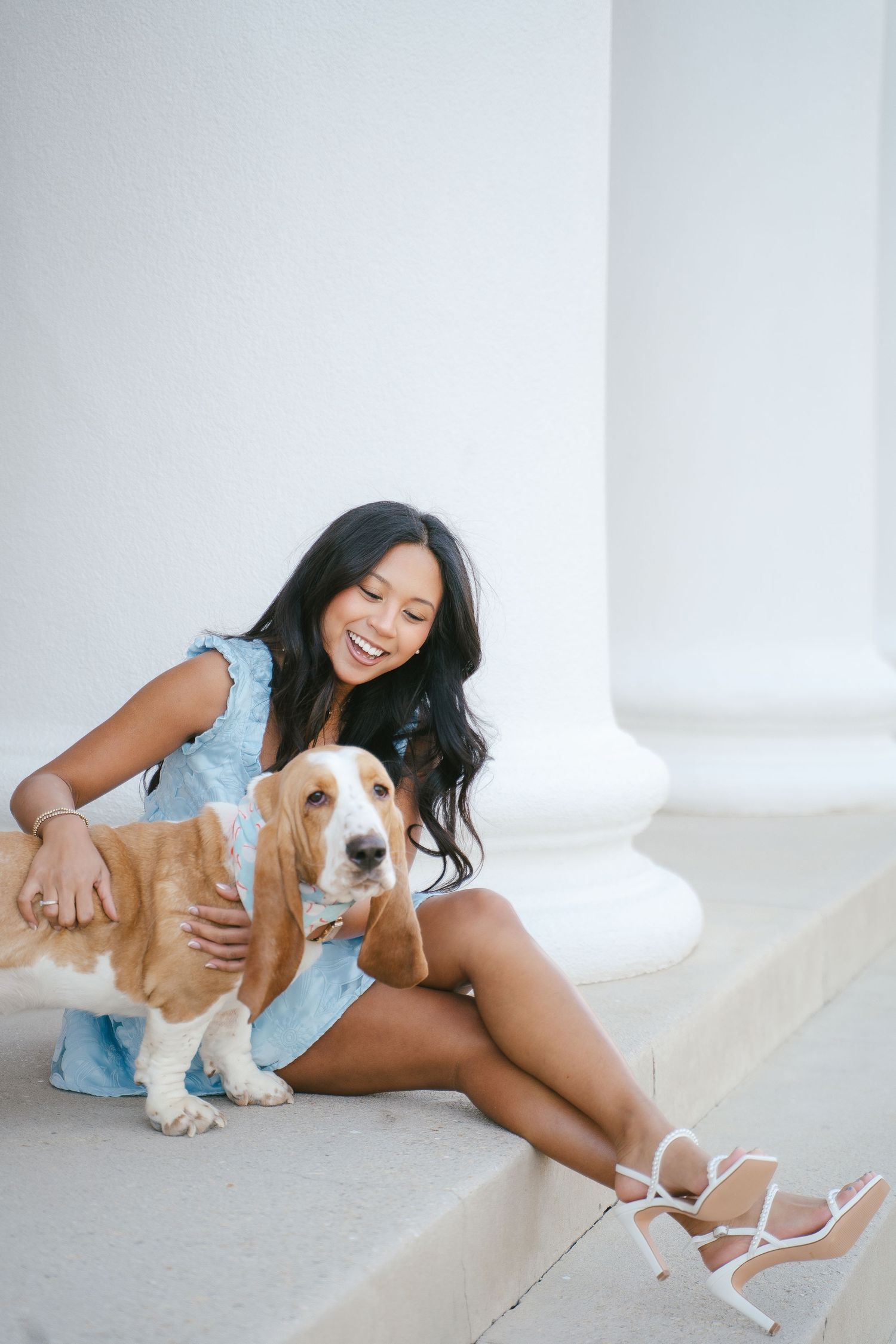 Person in blue dress sitting with Basset Hound dog against white wall.