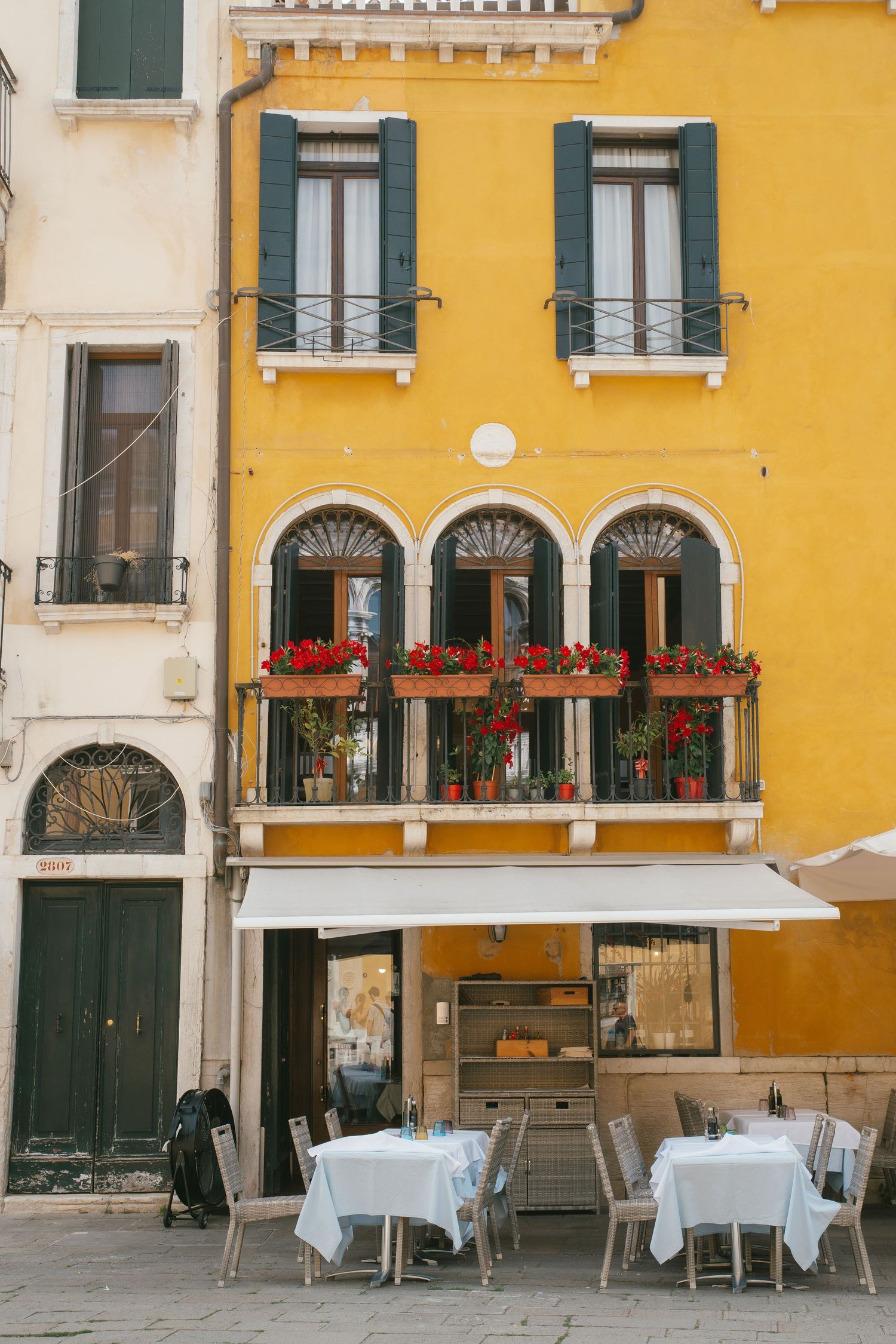 Yellow Italian building with green shutters and flower boxes on balcony above outdoor cafe tables.