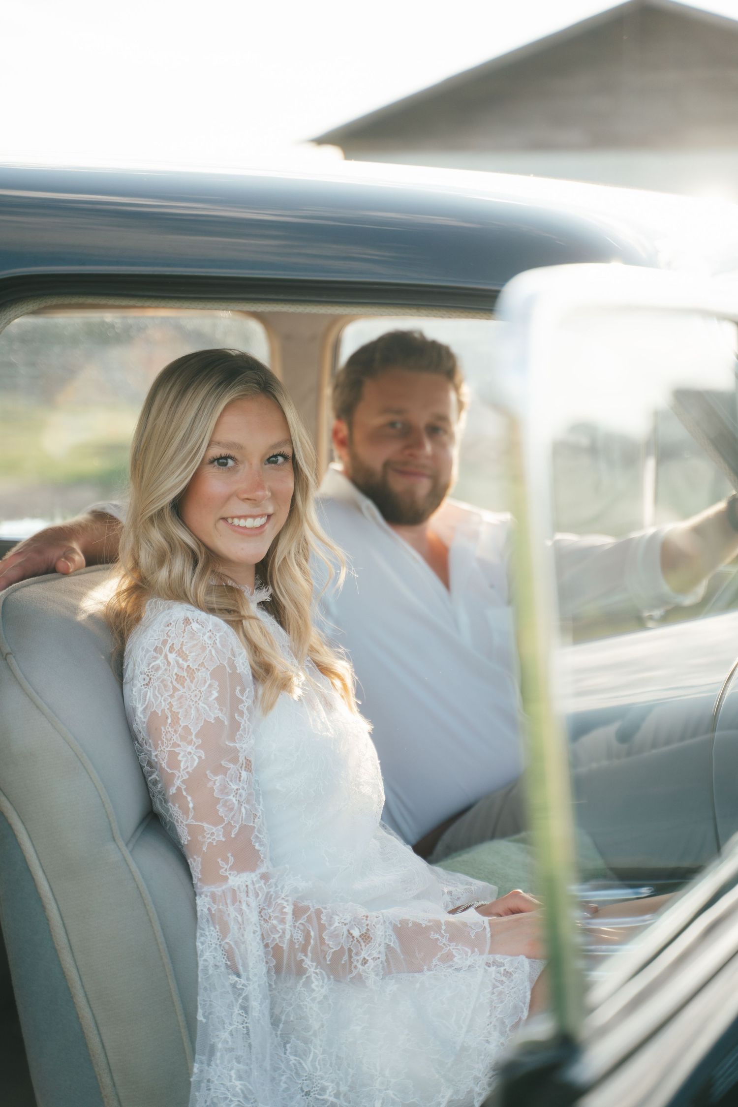 Smiling bride in white lace dress sits with groom in vintage car on their wedding day.