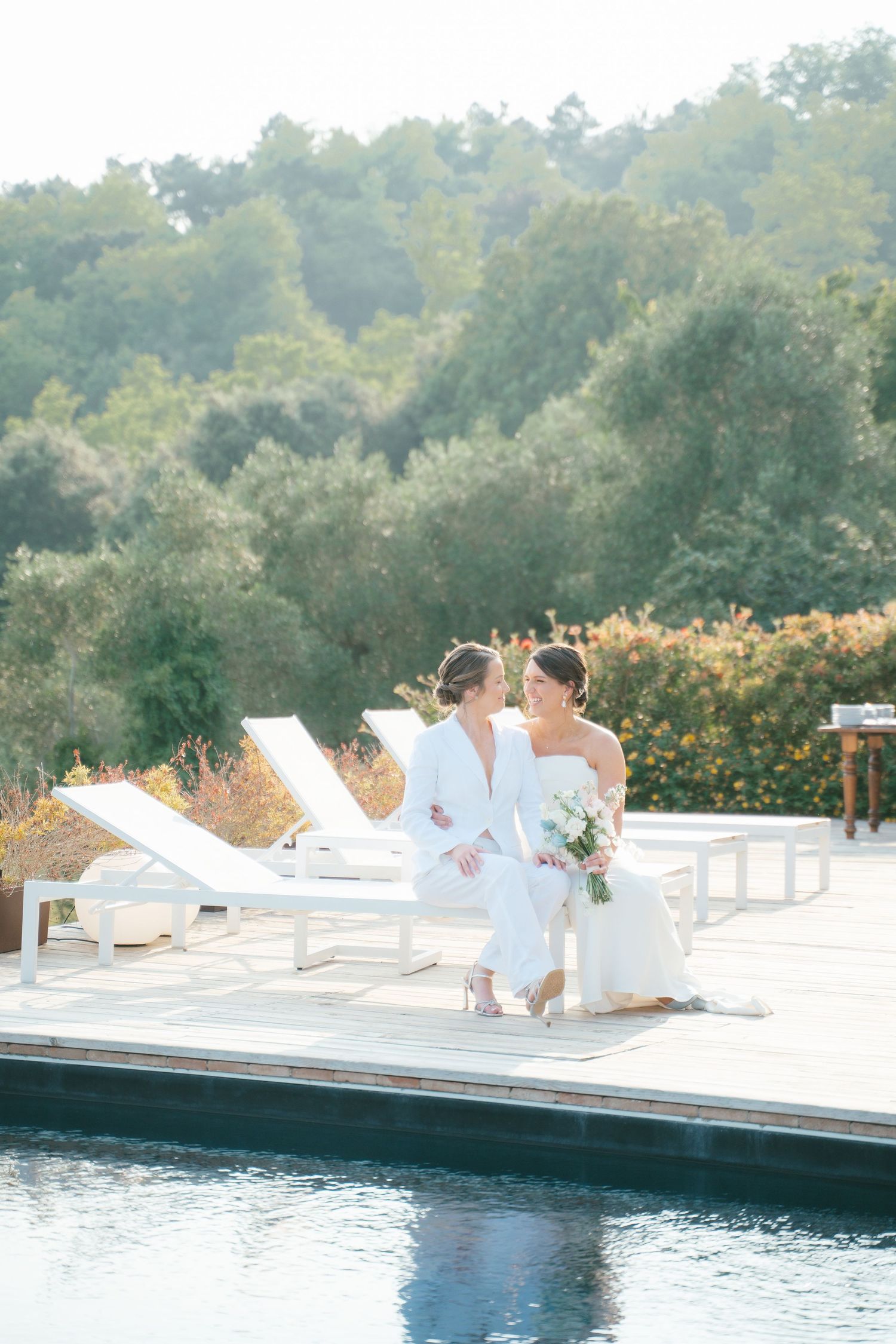 Two people in white clothing relaxing on pool deck chairs against a mountainous backdrop.