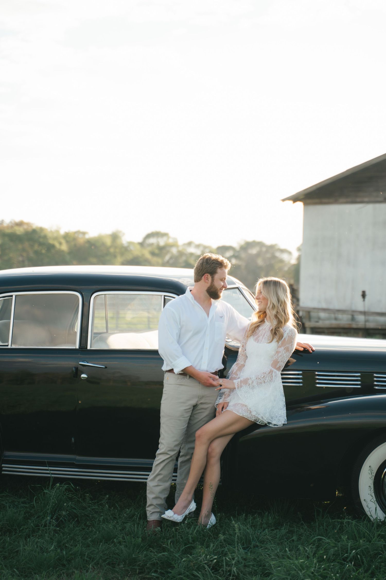 Couple posing romantically against a vintage black car in a rustic field at golden hour, woman in white lace dress.