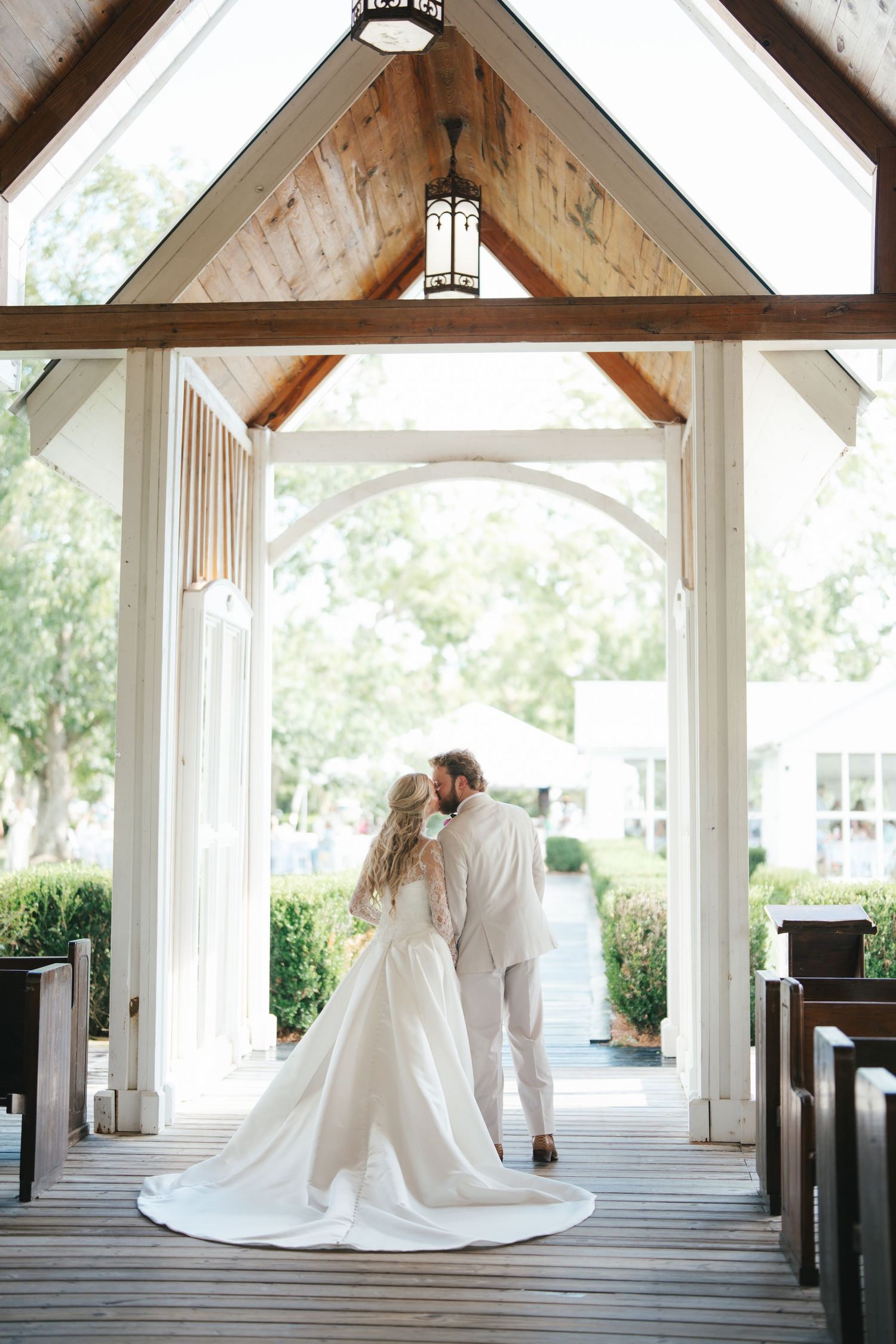 Bride and groom share a romantic kiss at the entrance of a white chapel with wooden beams and arched doors.