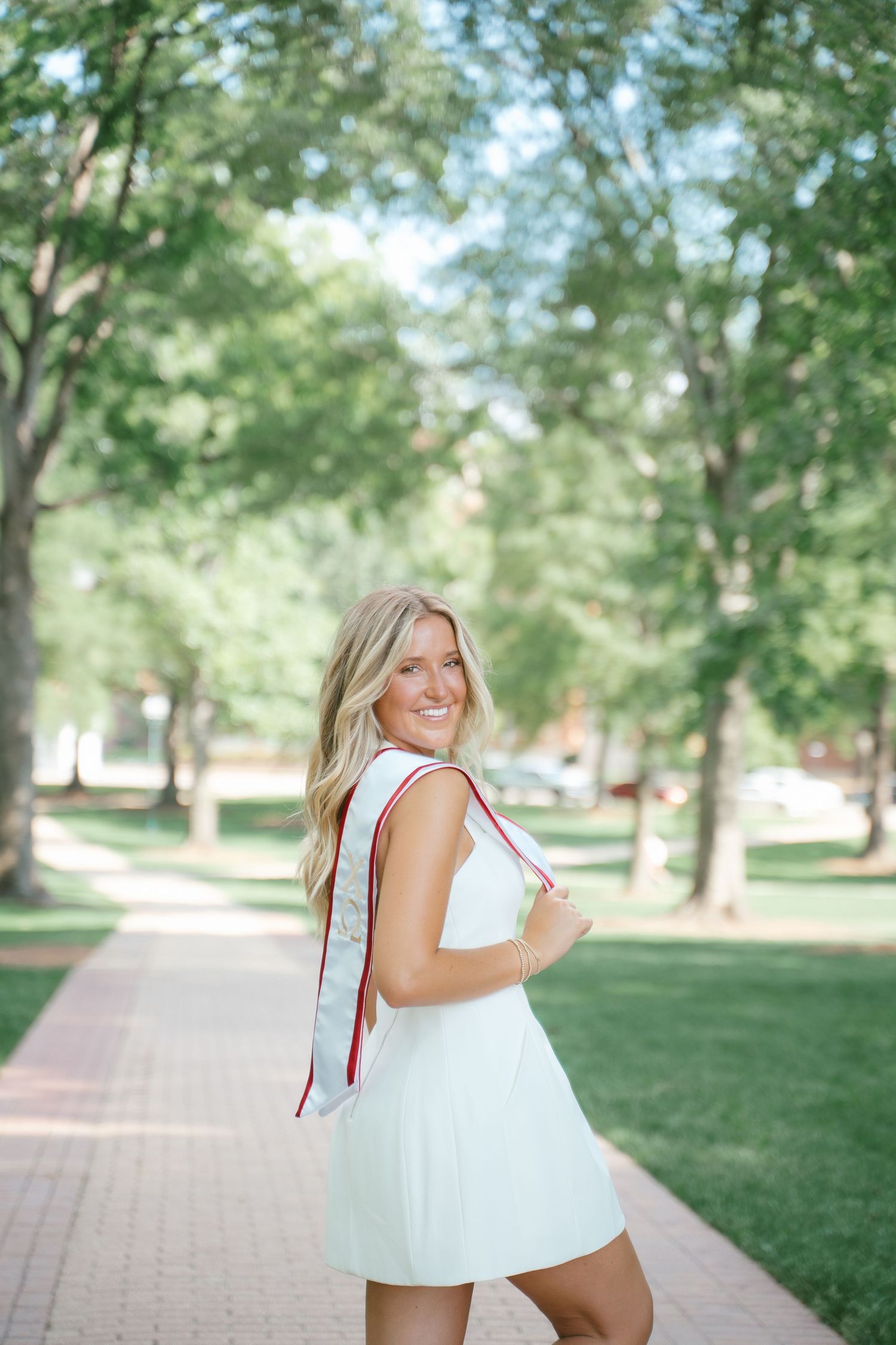 Person in white dress walking along tree-lined campus pathway.