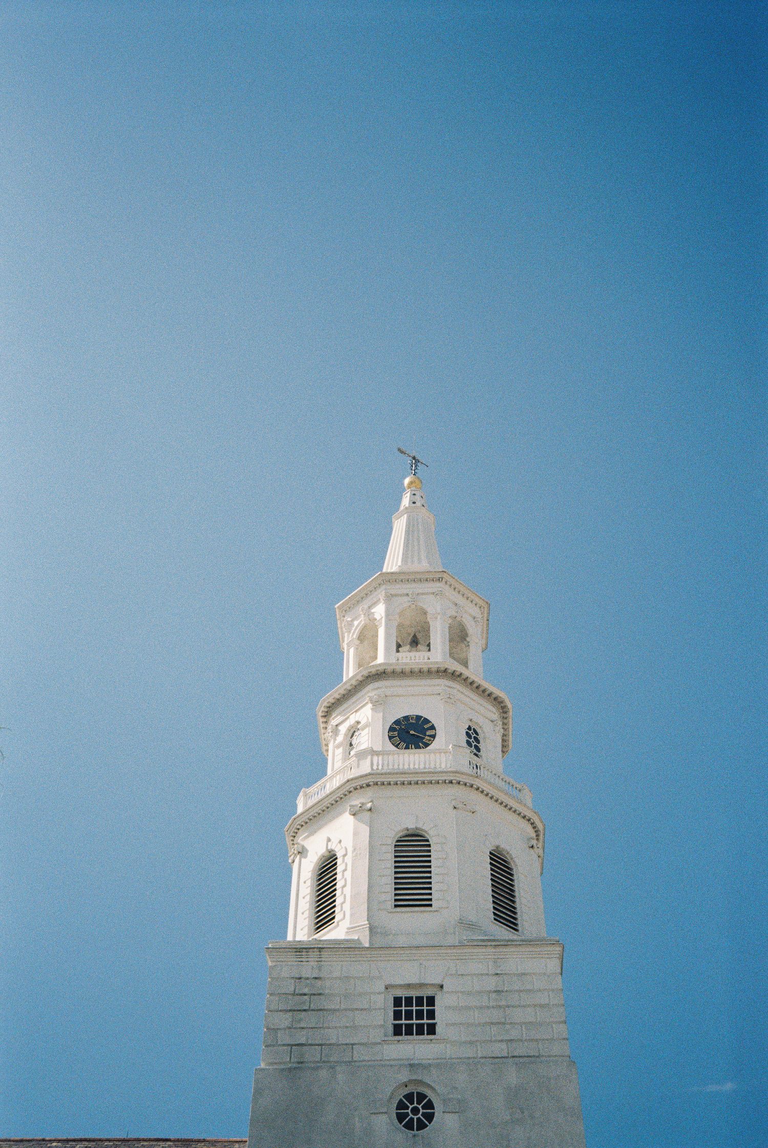 White church steeple with clock and weather vane against clear blue sky.