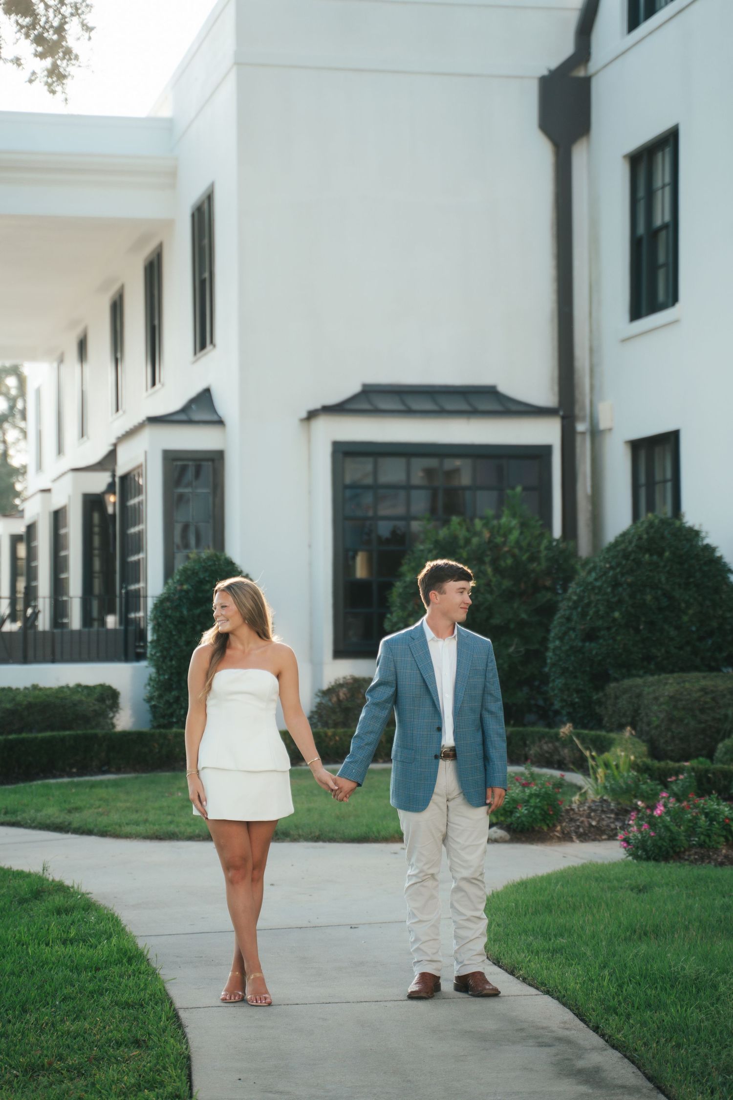Couple holding hands on a pathway outside a white estate, woman in white mini dress, man in blue blazer and khakis.