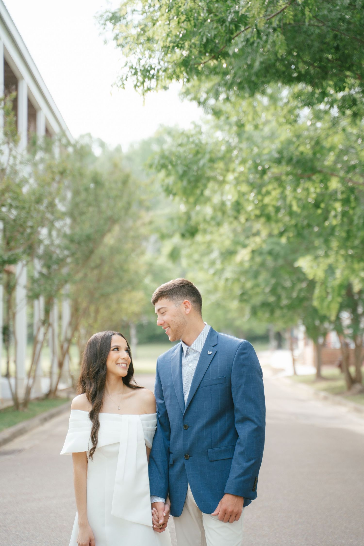 A couple in formal and casual wear strolls down a tree-lined path.
