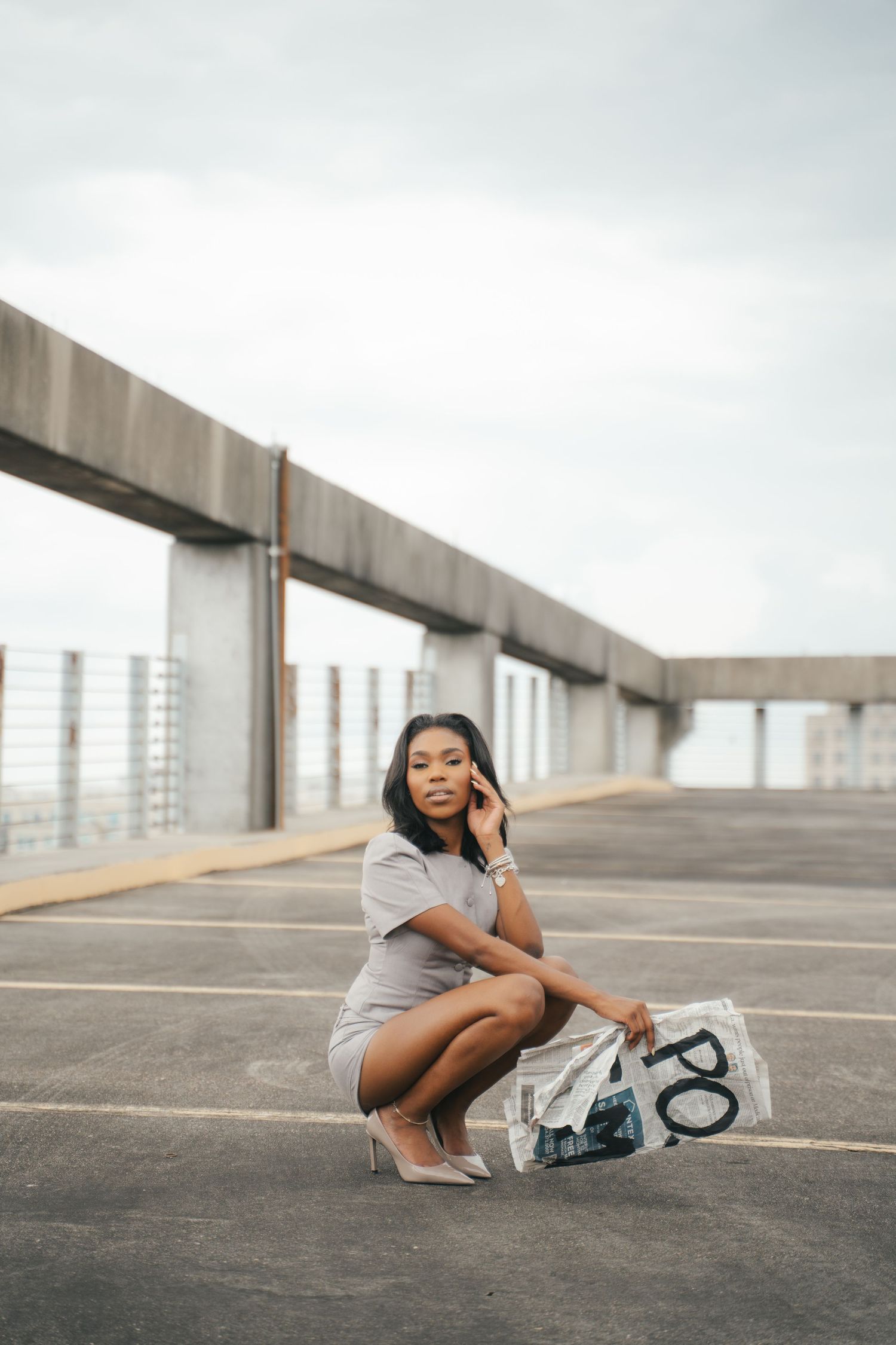 Young Black woman in gray dress crouching on rooftop parking lot, holding newspaper, wearing heels.