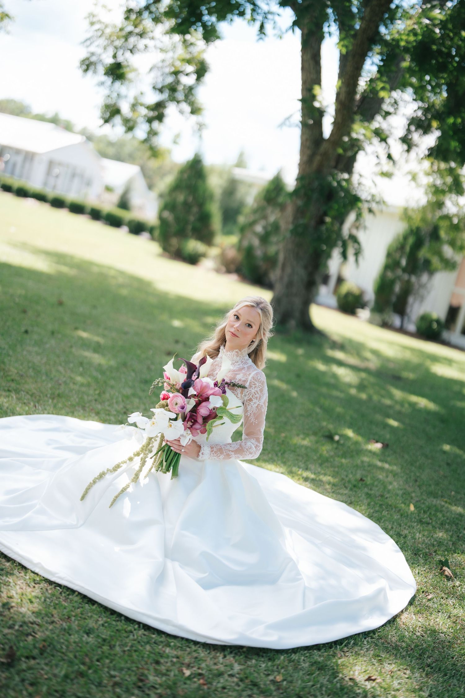 Bride in lace long-sleeve wedding gown holding floral bouquet on green lawn with large tree backdrop.
