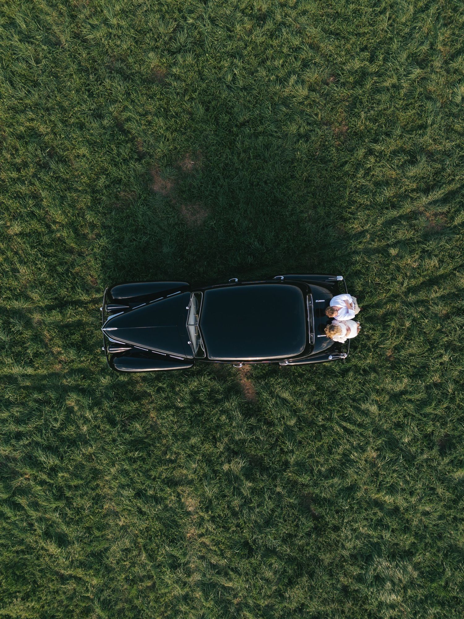 Aerial drone view of a classic black vintage car parked in a green field with two people visible inside.