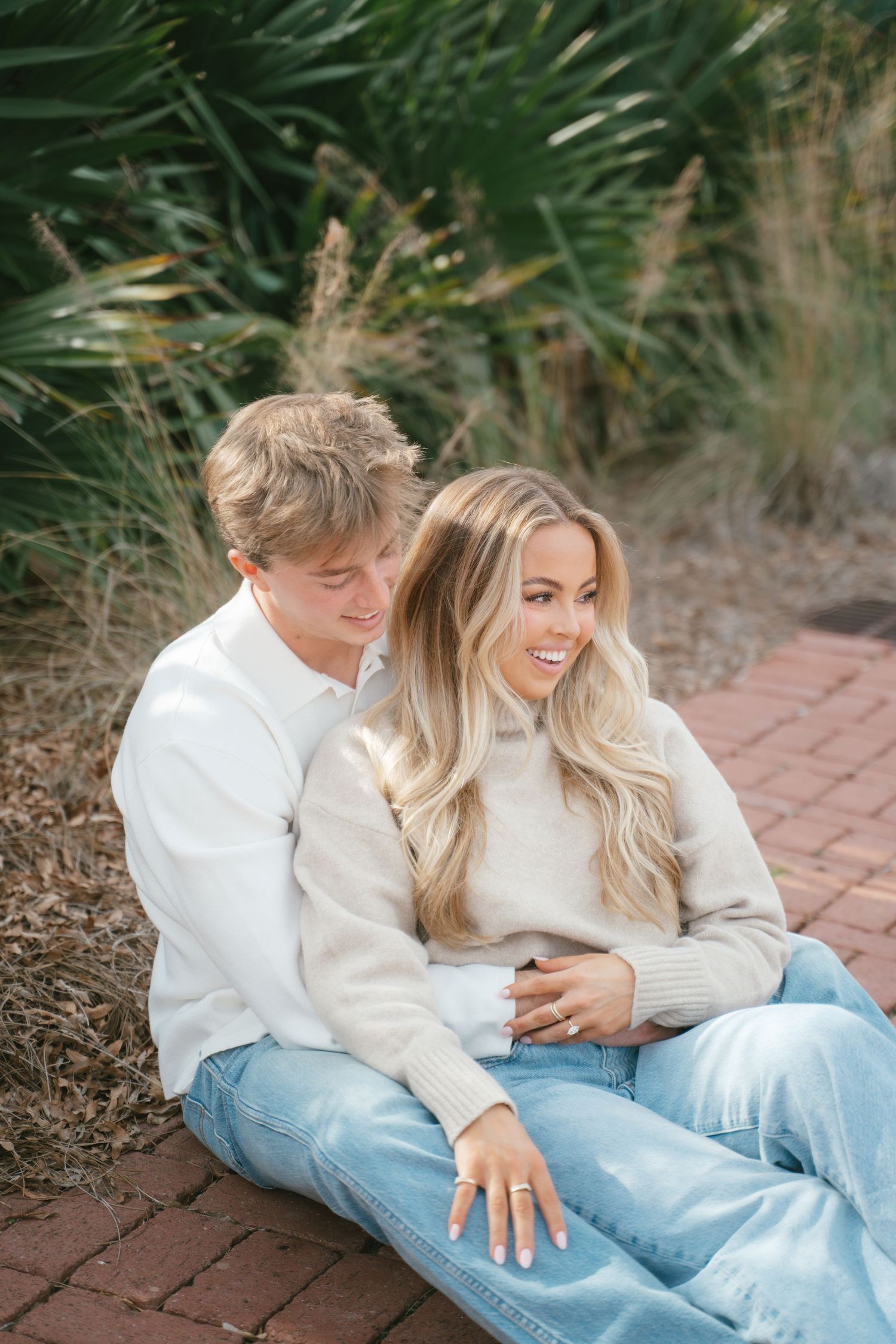 A couple in matching white sweaters and jeans sits together on brick pavement.