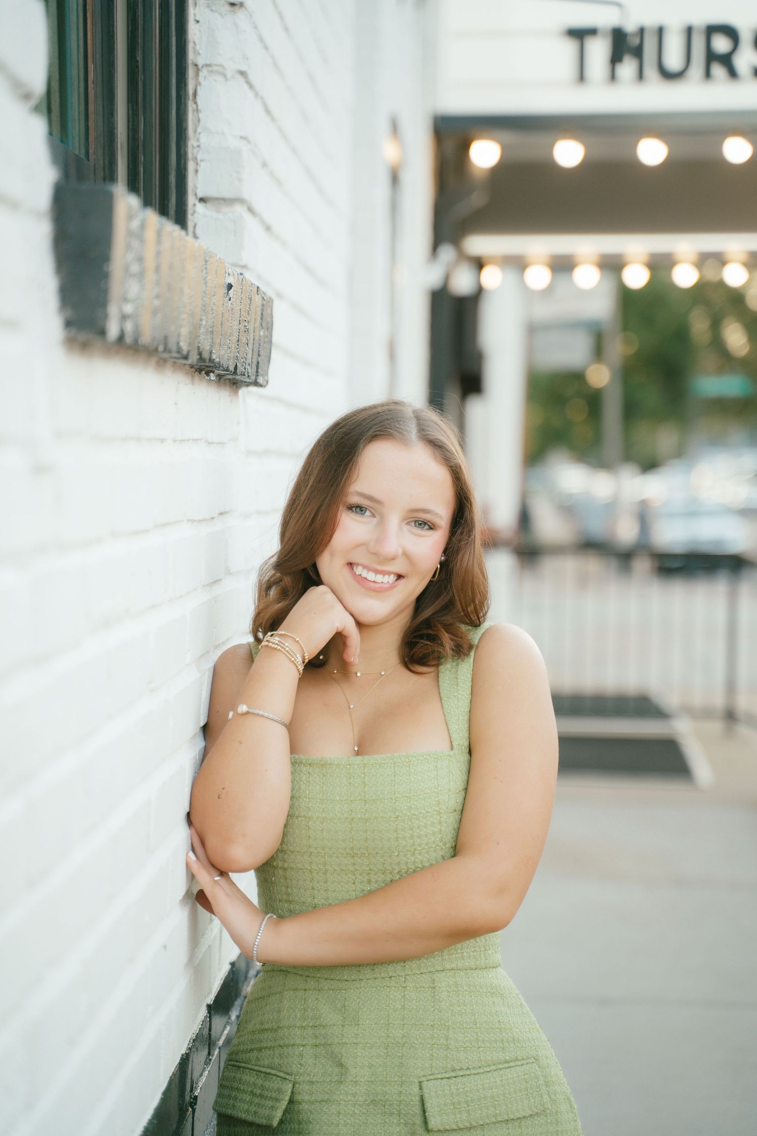 Person in green top posing against white wall with string lights.