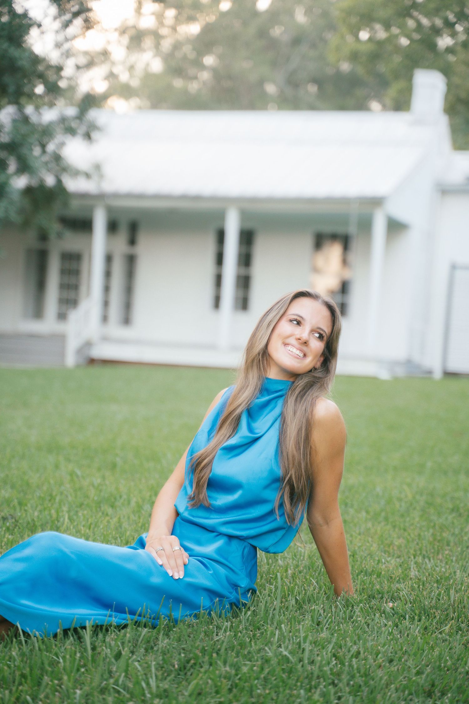 Casual pose on grass wearing vibrant blue dress with white farmhouse in background.