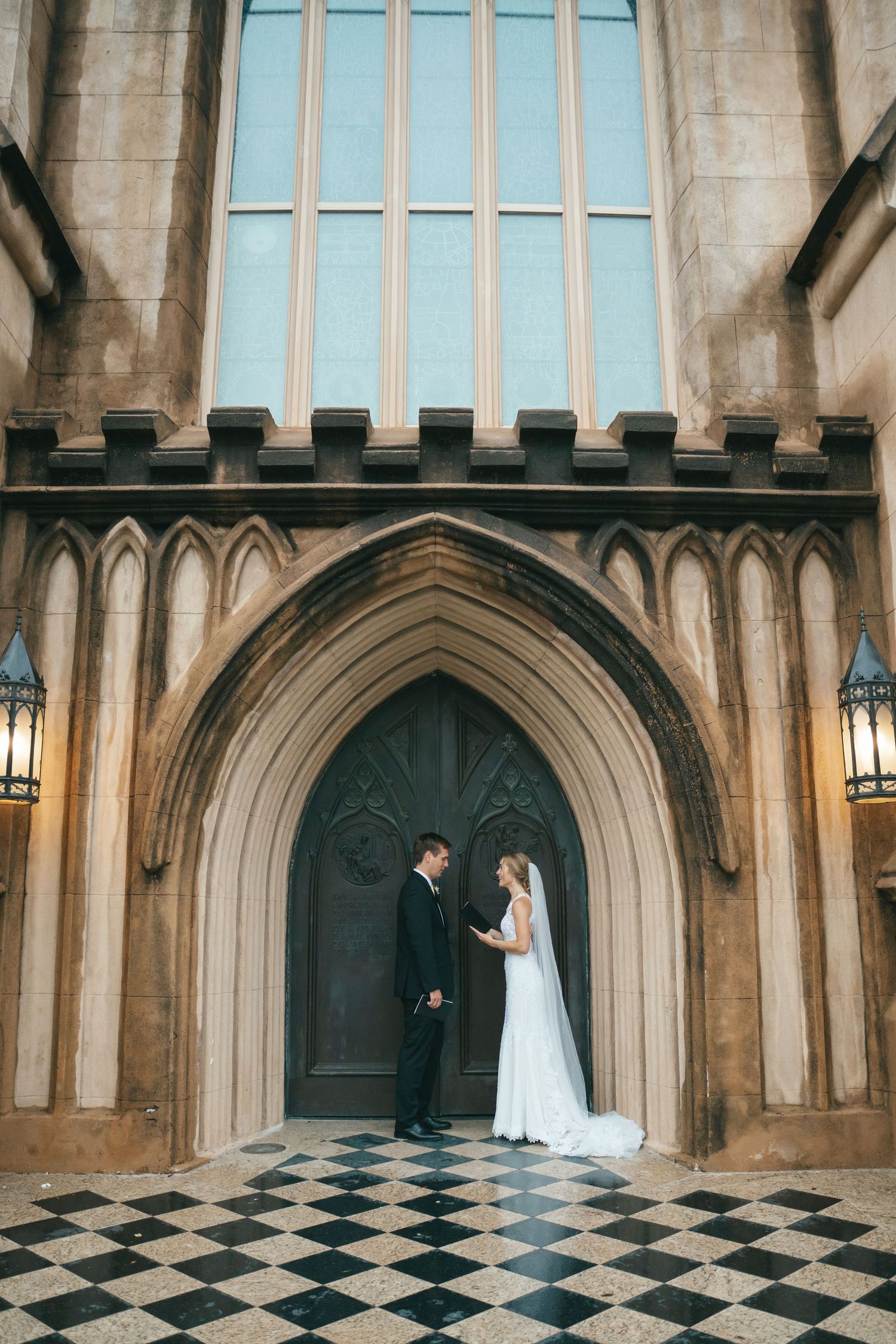 Bride and groom exchange vows at ornate Gothic church entrance with checkered floor and arched wooden doors.