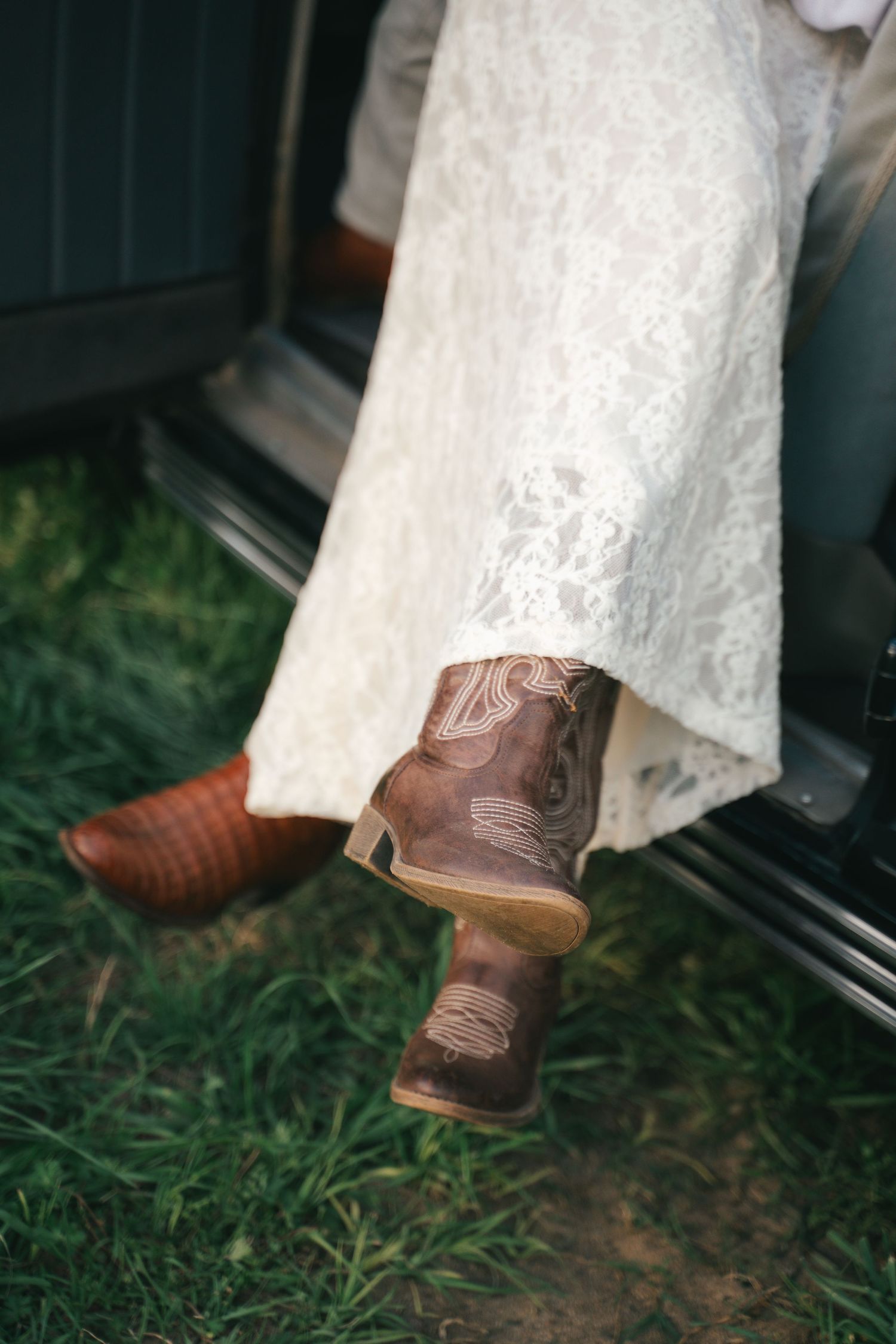 Bride in lace wedding dress wearing brown leather cowboy boots, sitting in a vintage vehicle on green grass.