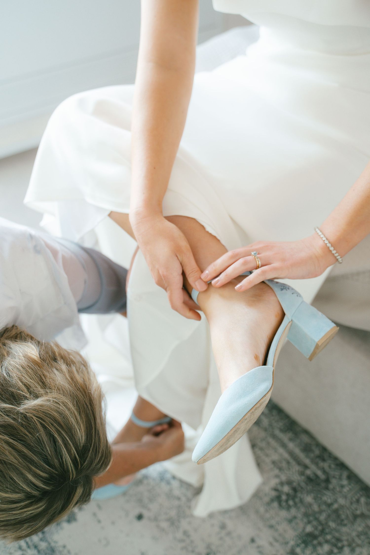 Someone helping a bride put on light blue wedding shoes while wearing a white dress.