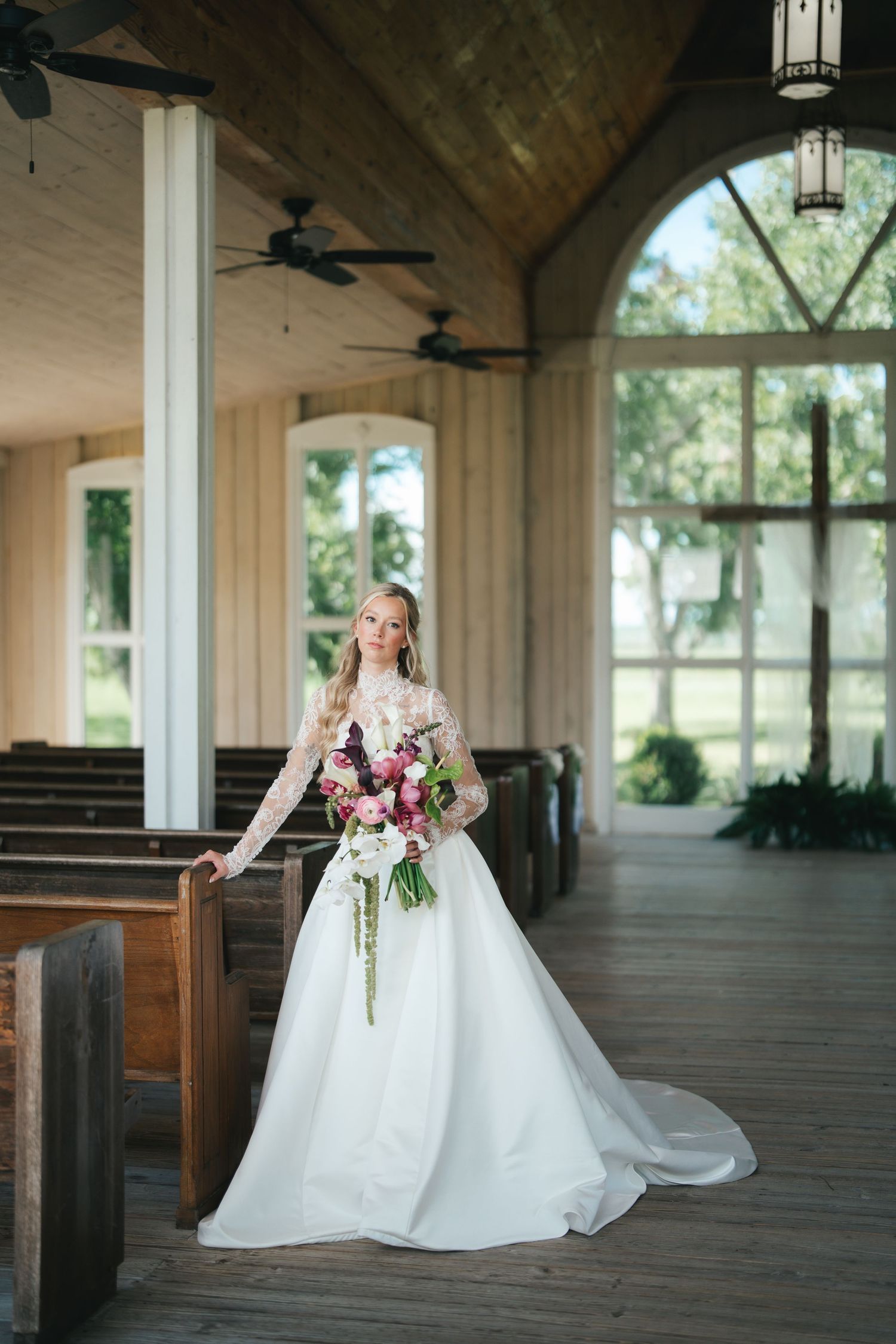 Bride in white lace long-sleeve gown holds floral bouquet inside elegant wood chapel with arched windows.