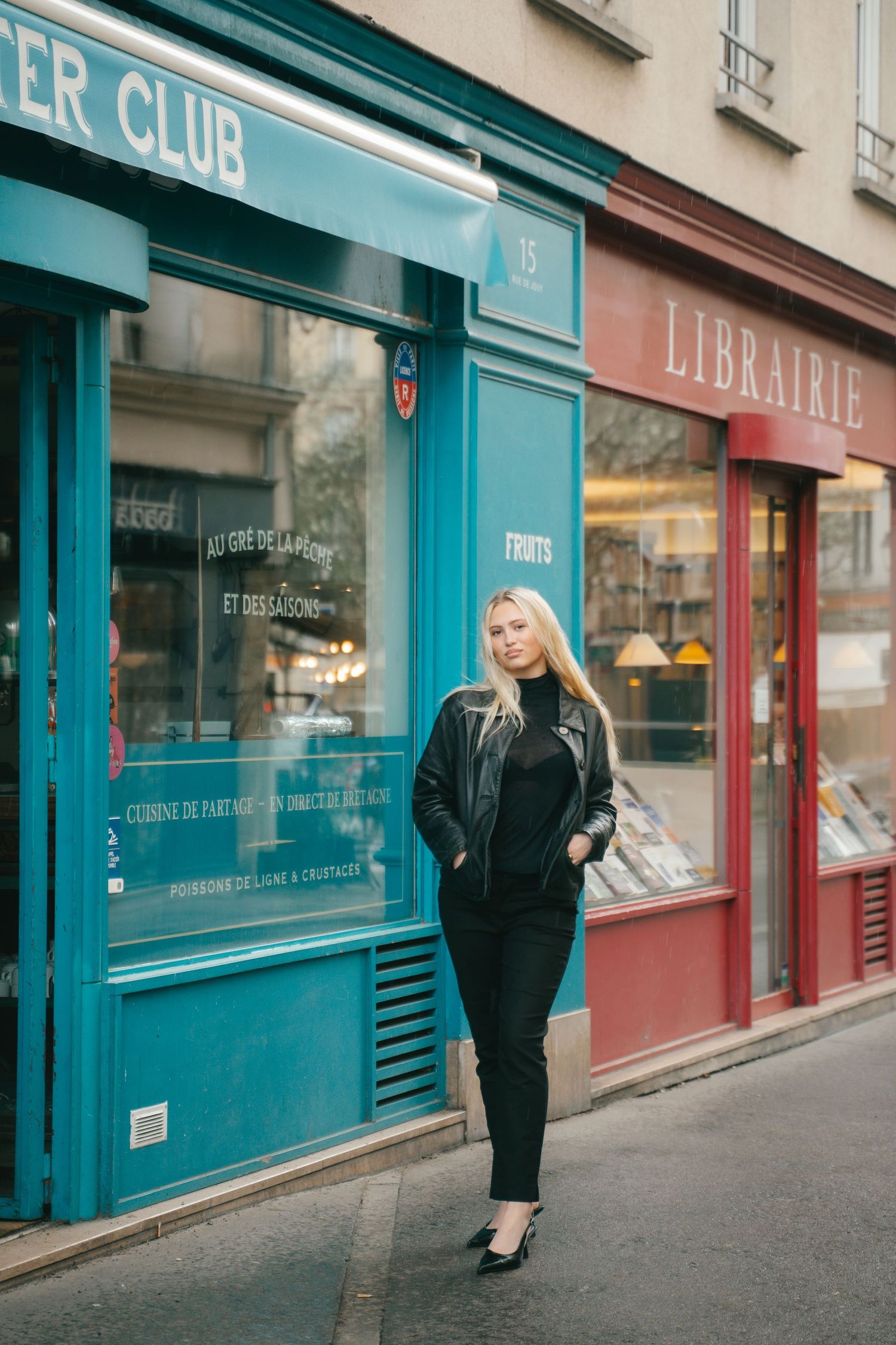 Blonde woman in black outfit posing on a Parisian street beside a teal shop and red librairie storefront.