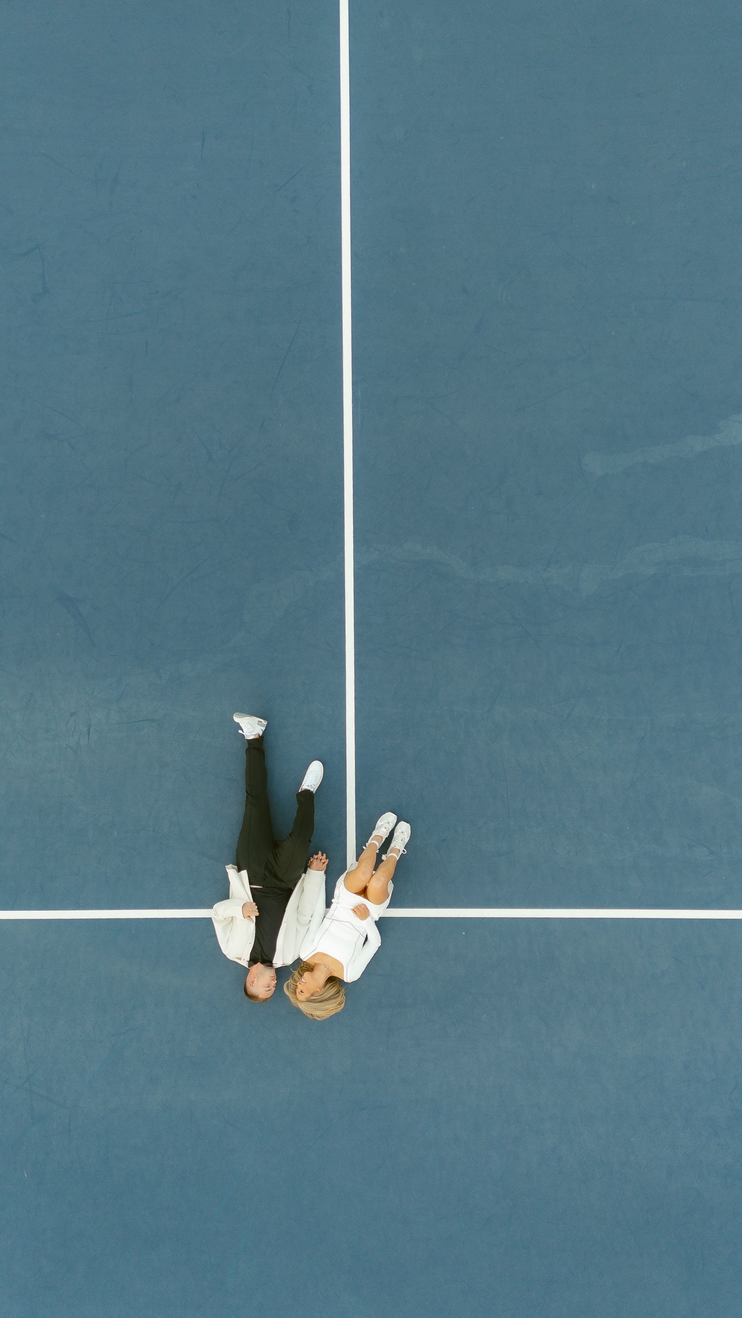 Aerial view of couple sitting together on blue tennis court surface.