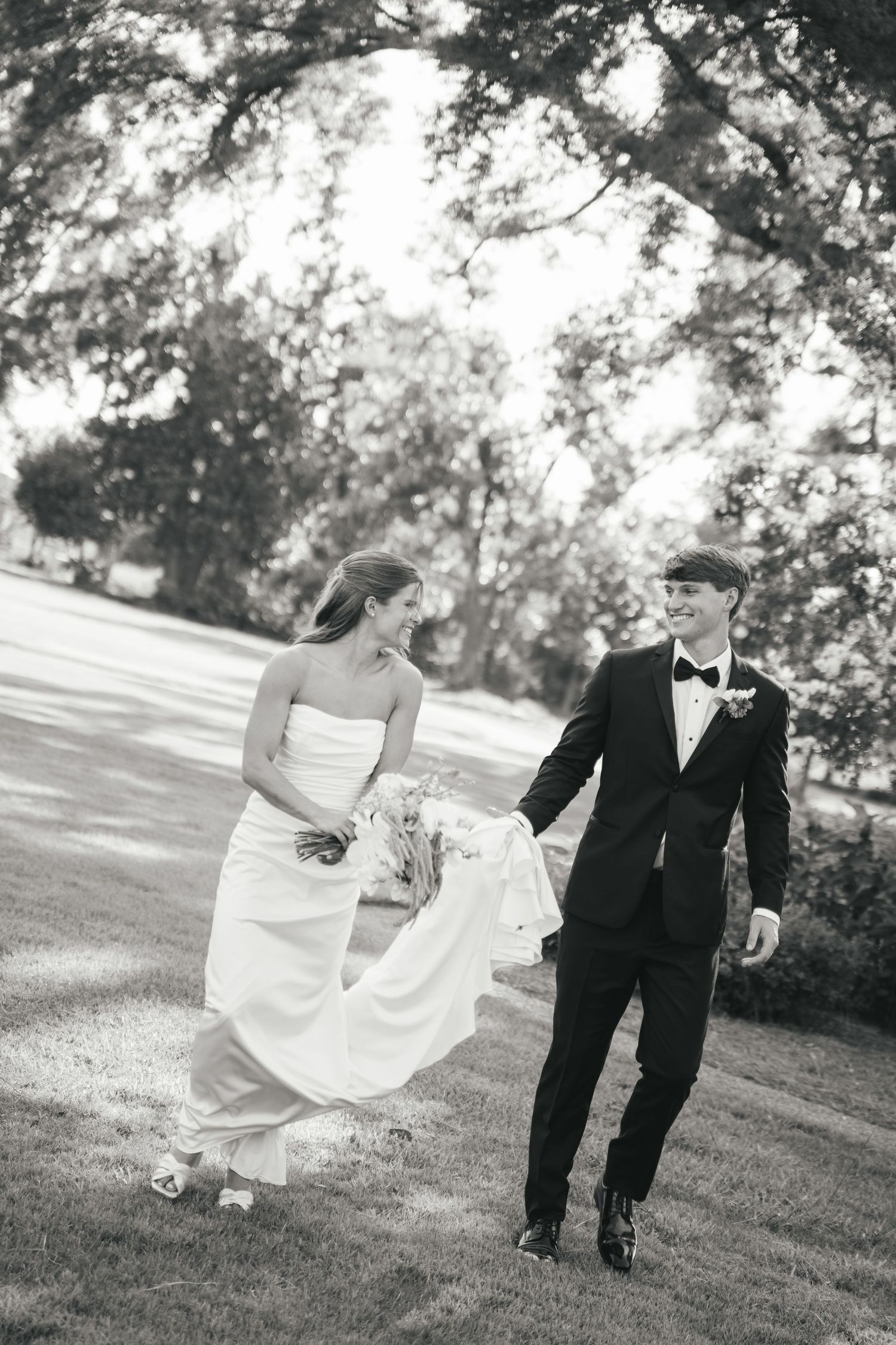 Black and white photo of a smiling bride and groom walking hand in hand outdoors under large oak trees.