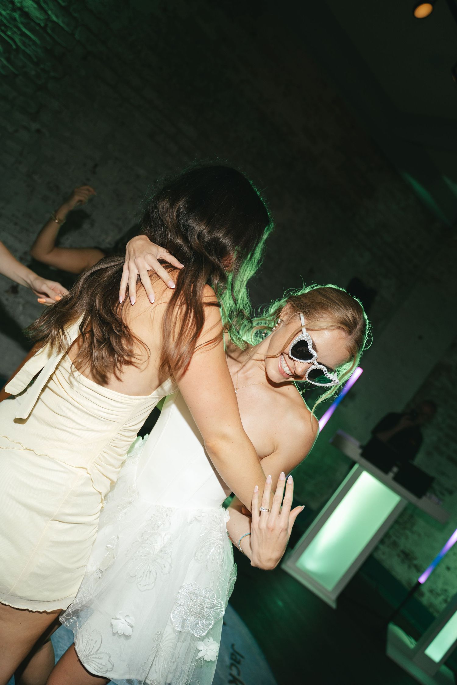 Two women dancing together at a party, one in white dress with green hair and heart-shaped sunglasses under green lights.