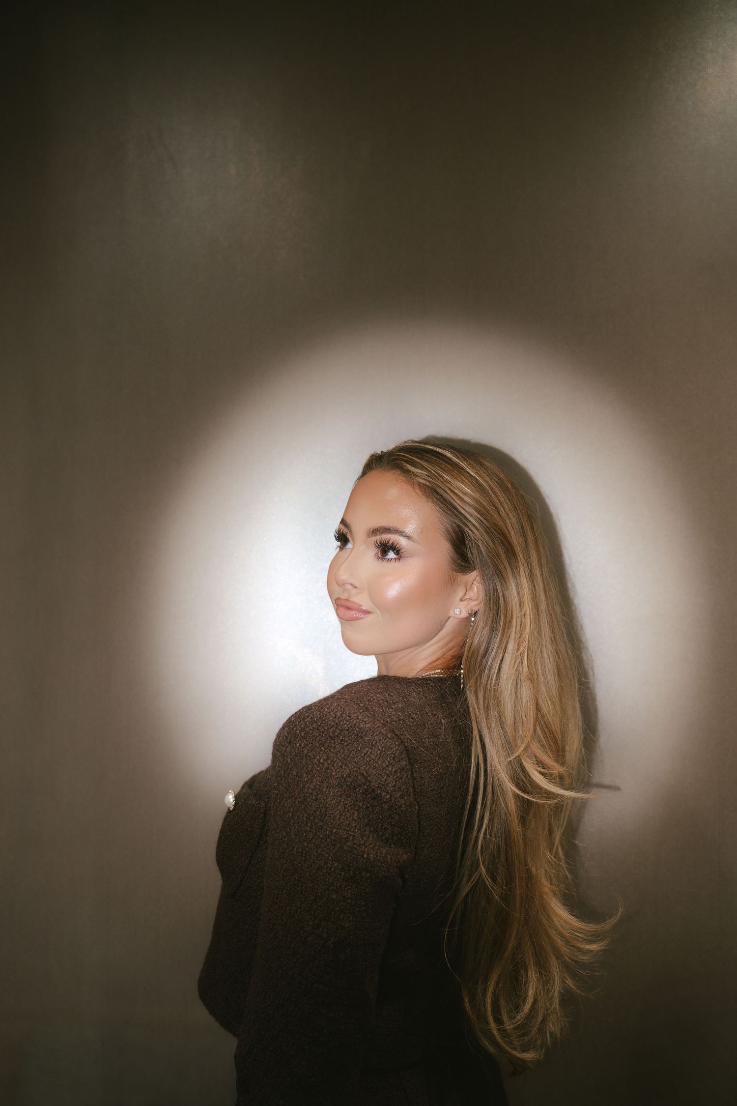 Young woman with long blonde hair in dark top poses against illuminated wall in dramatic portrait.