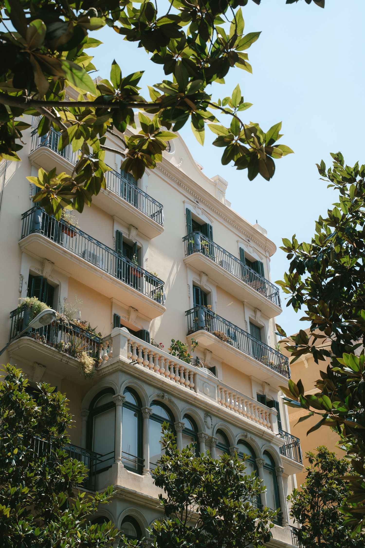 Mediterranean style apartment building with white facade and green balconies viewed through lush foliage.
