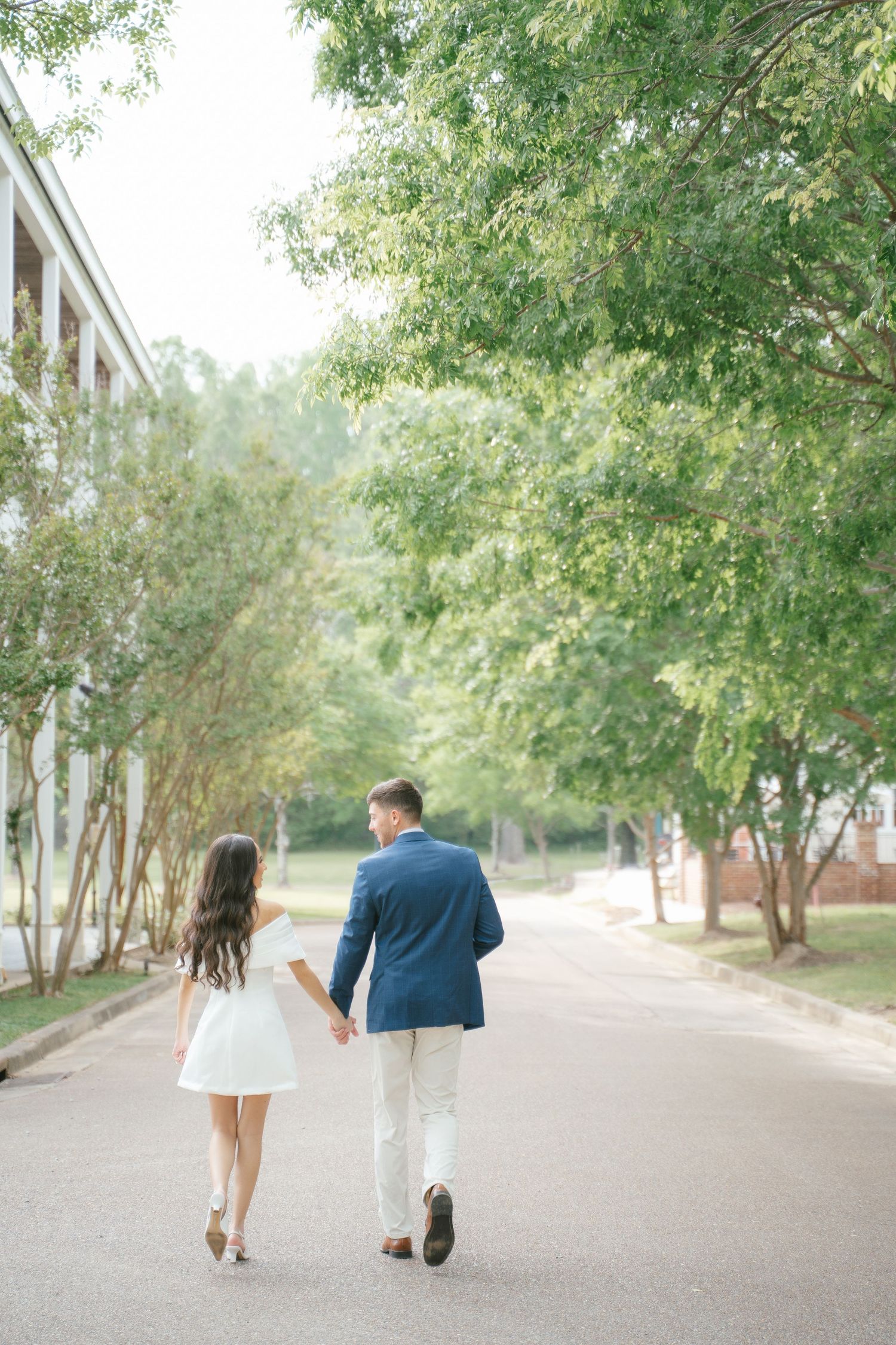 A couple walks hand in hand down a tree-lined path in elegant attire.