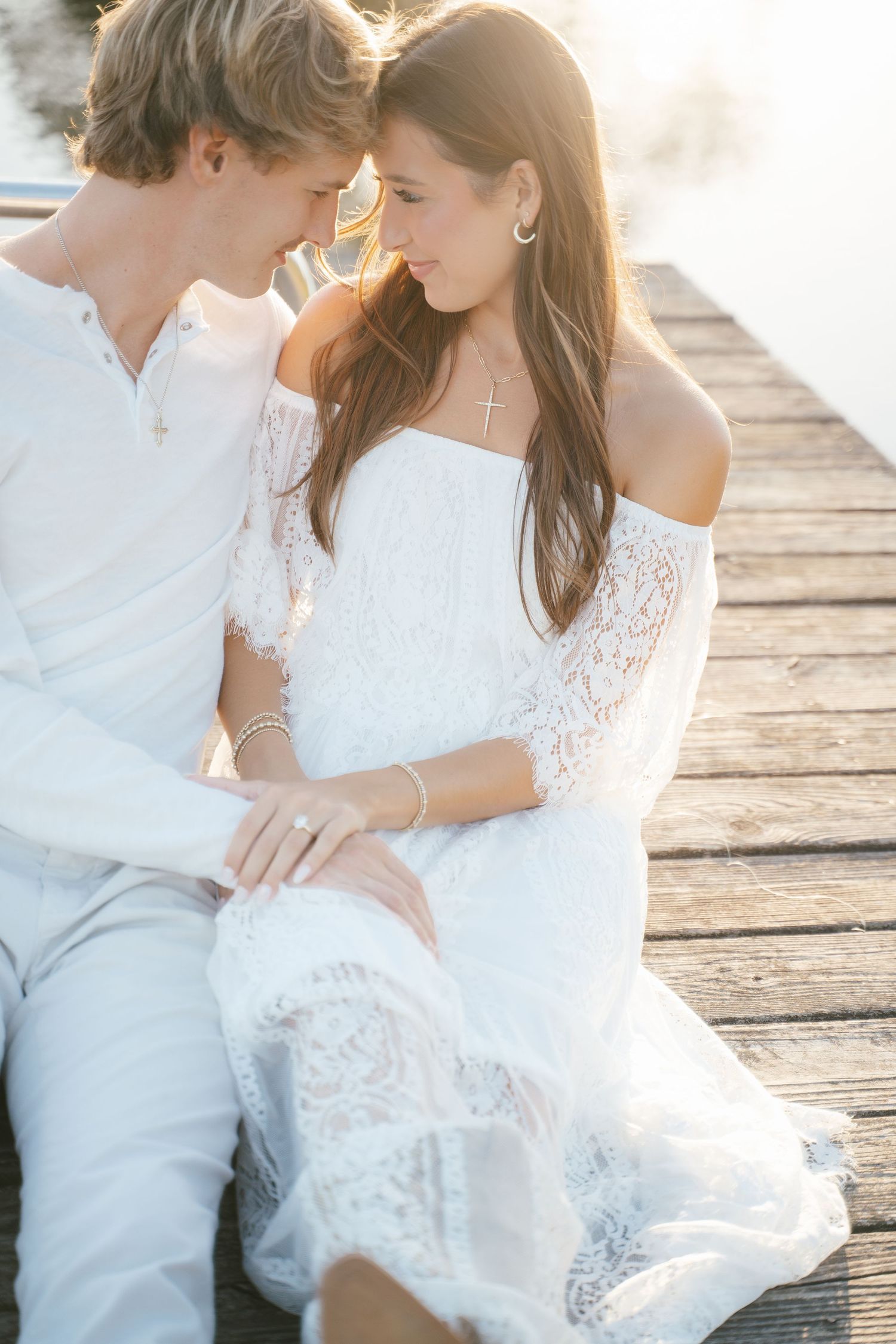 A couple in white attire shares tender moments on a wooden dock at sunset.
