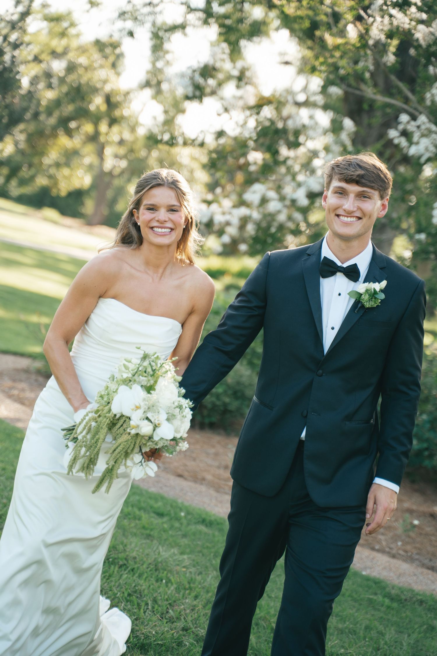Smiling bride in strapless white gown holding bouquet walks with groom in black tuxedo outdoors.