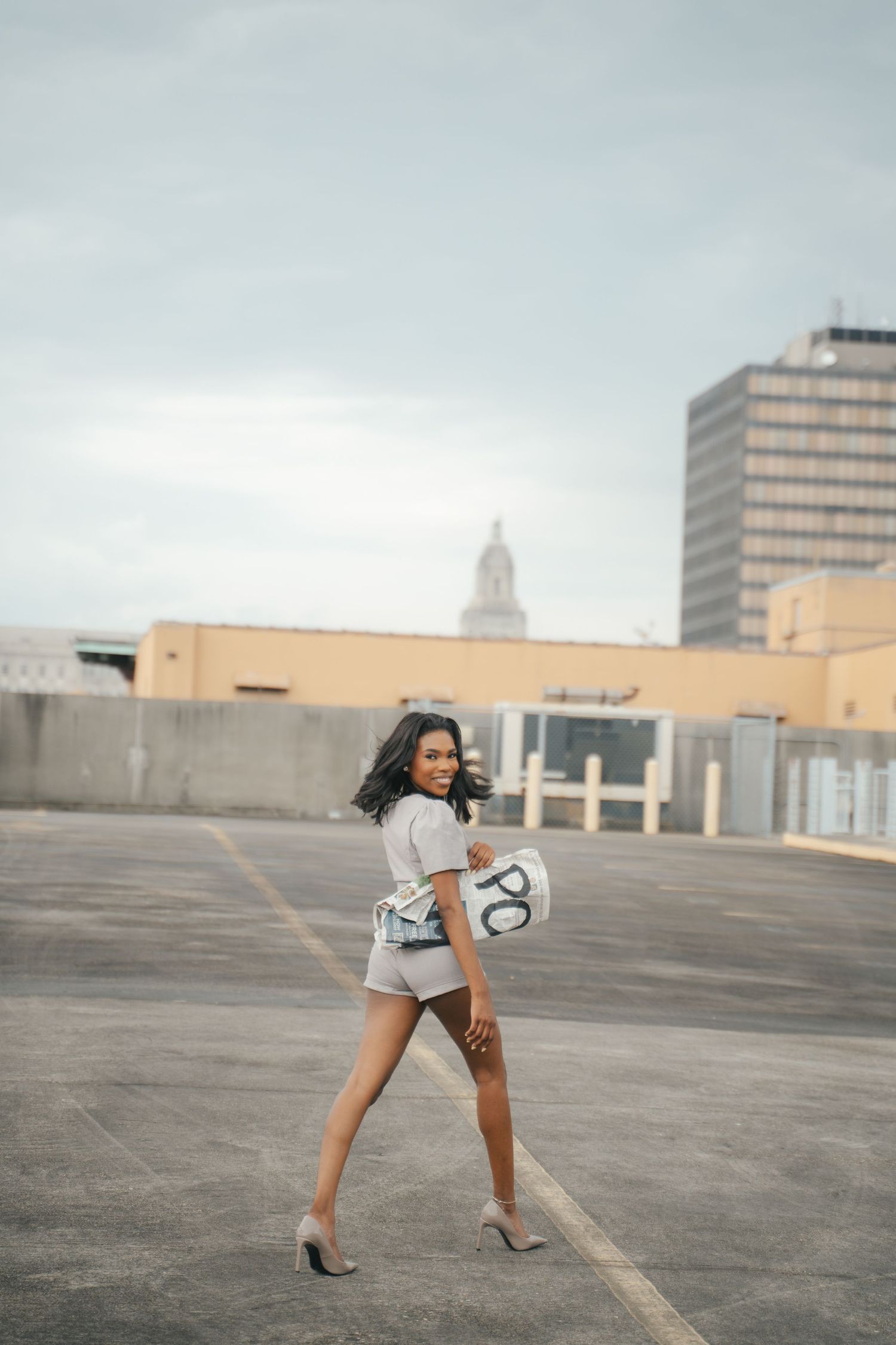 Young woman in gray romper and heels poses on rooftop parking lot with city buildings in background.