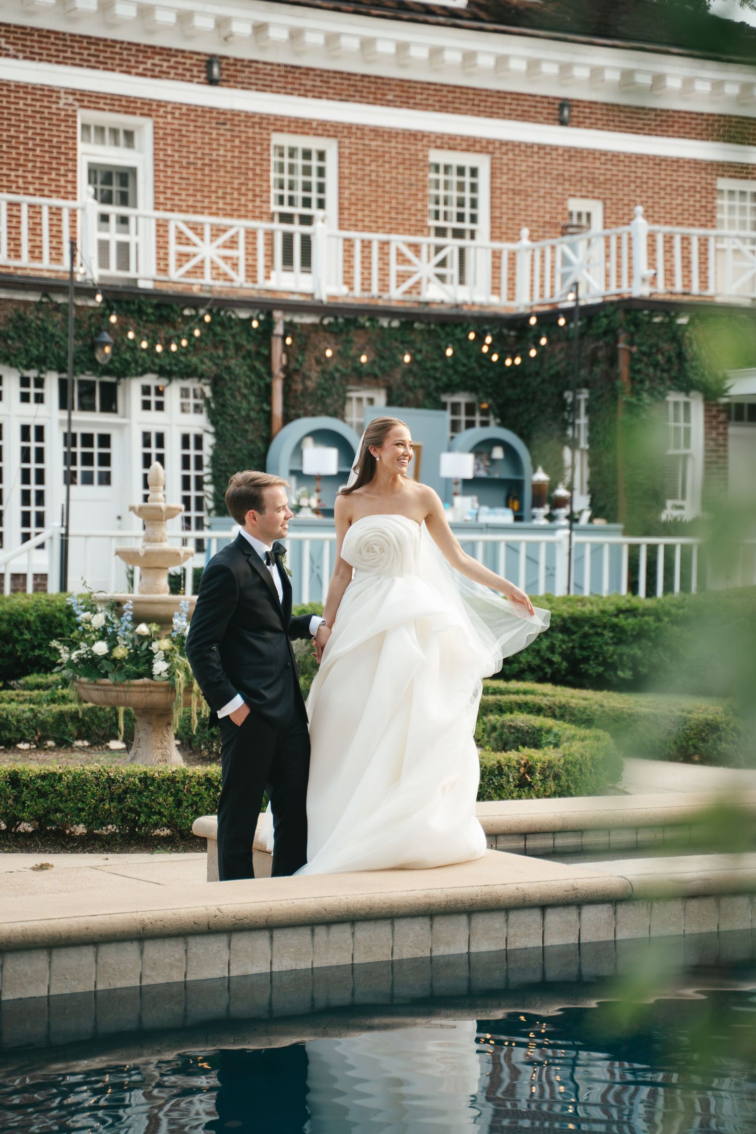 Bride in white strapless gown and groom in black tuxedo stand by a pool at an elegant brick estate wedding venue.