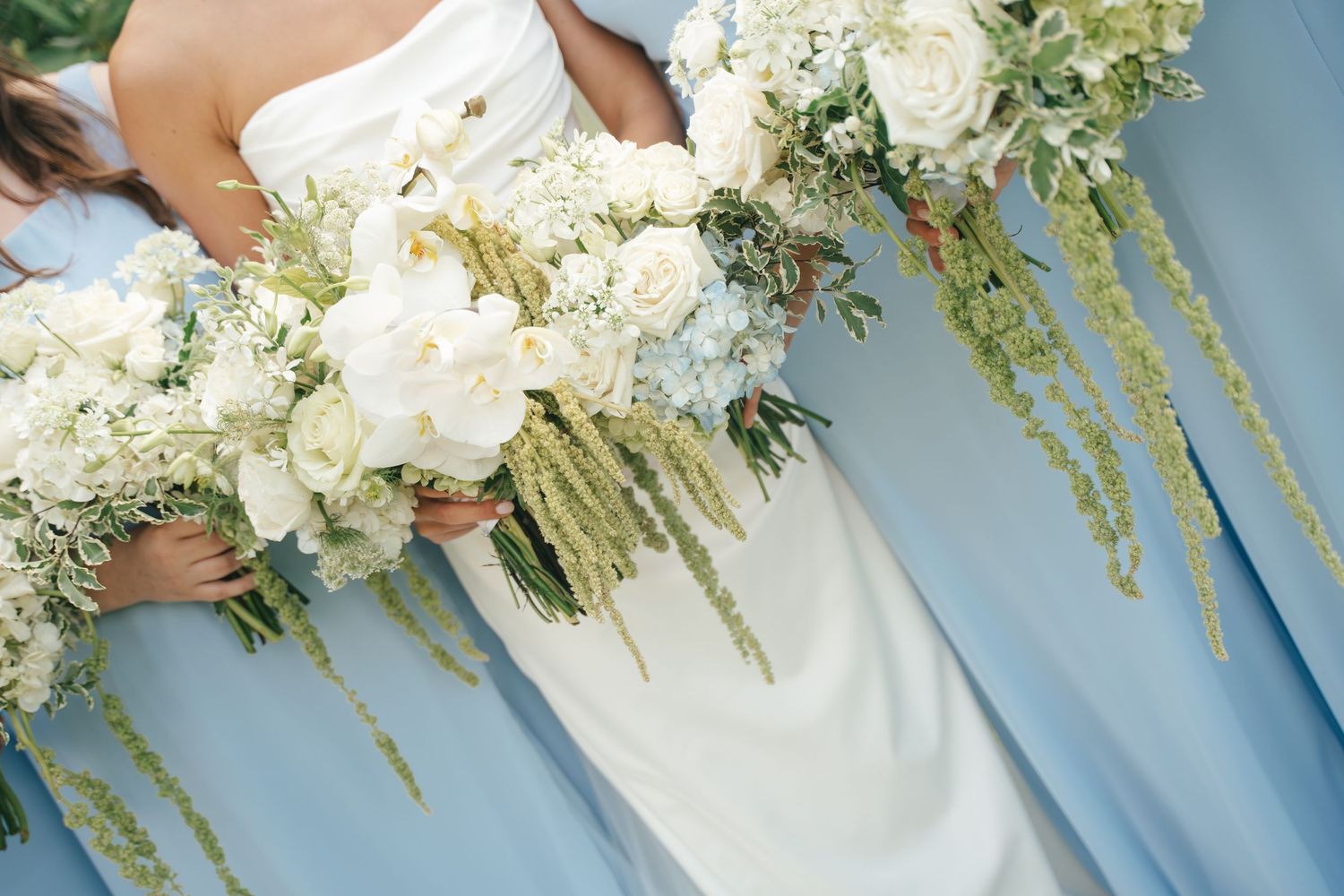 Bride and bridesmaids holding lush white floral bouquets with cascading greenery against soft blue dresses.