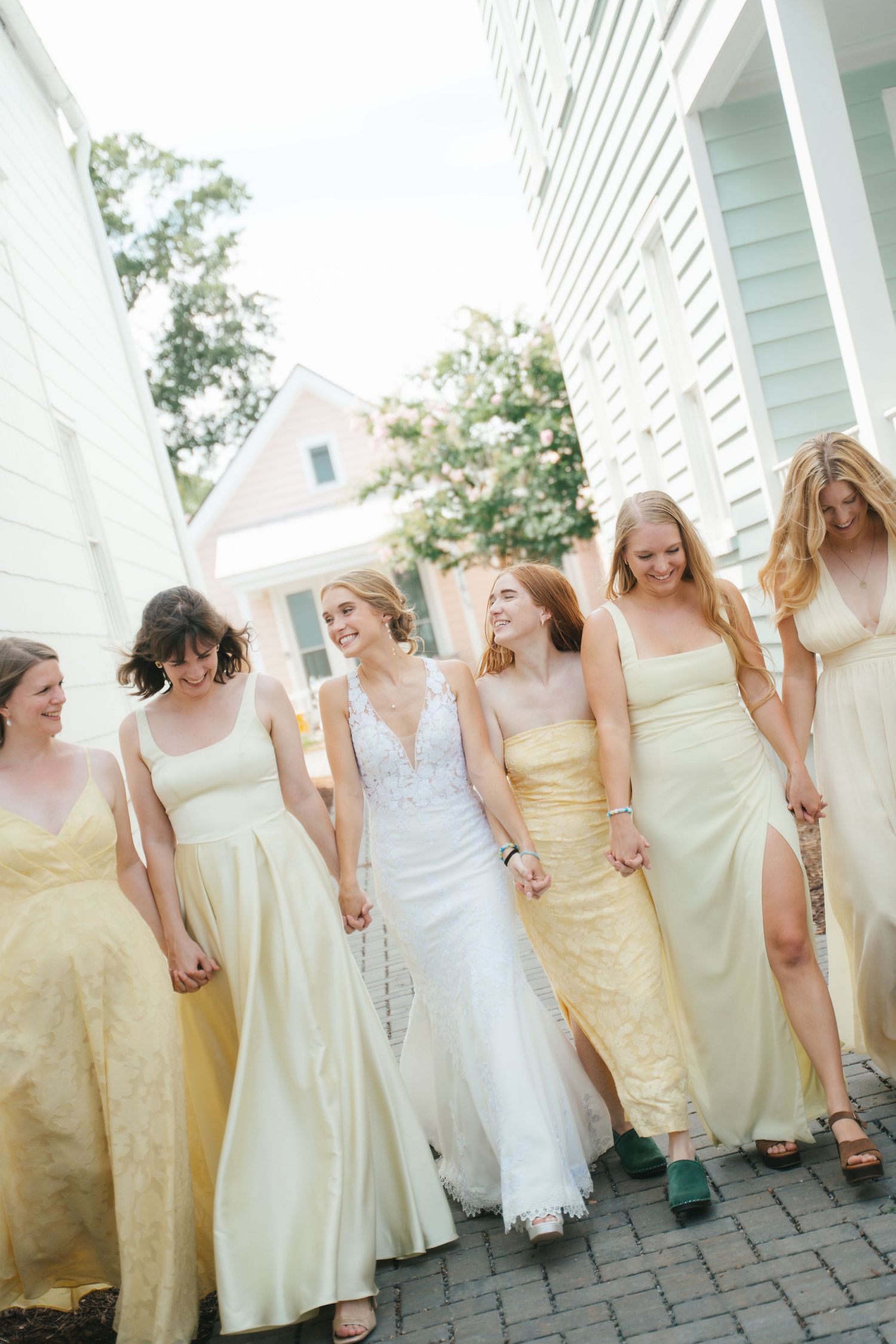 Bride in white lace gown walks with six bridesmaids in yellow dresses, holding hands outdoors near white buildings.